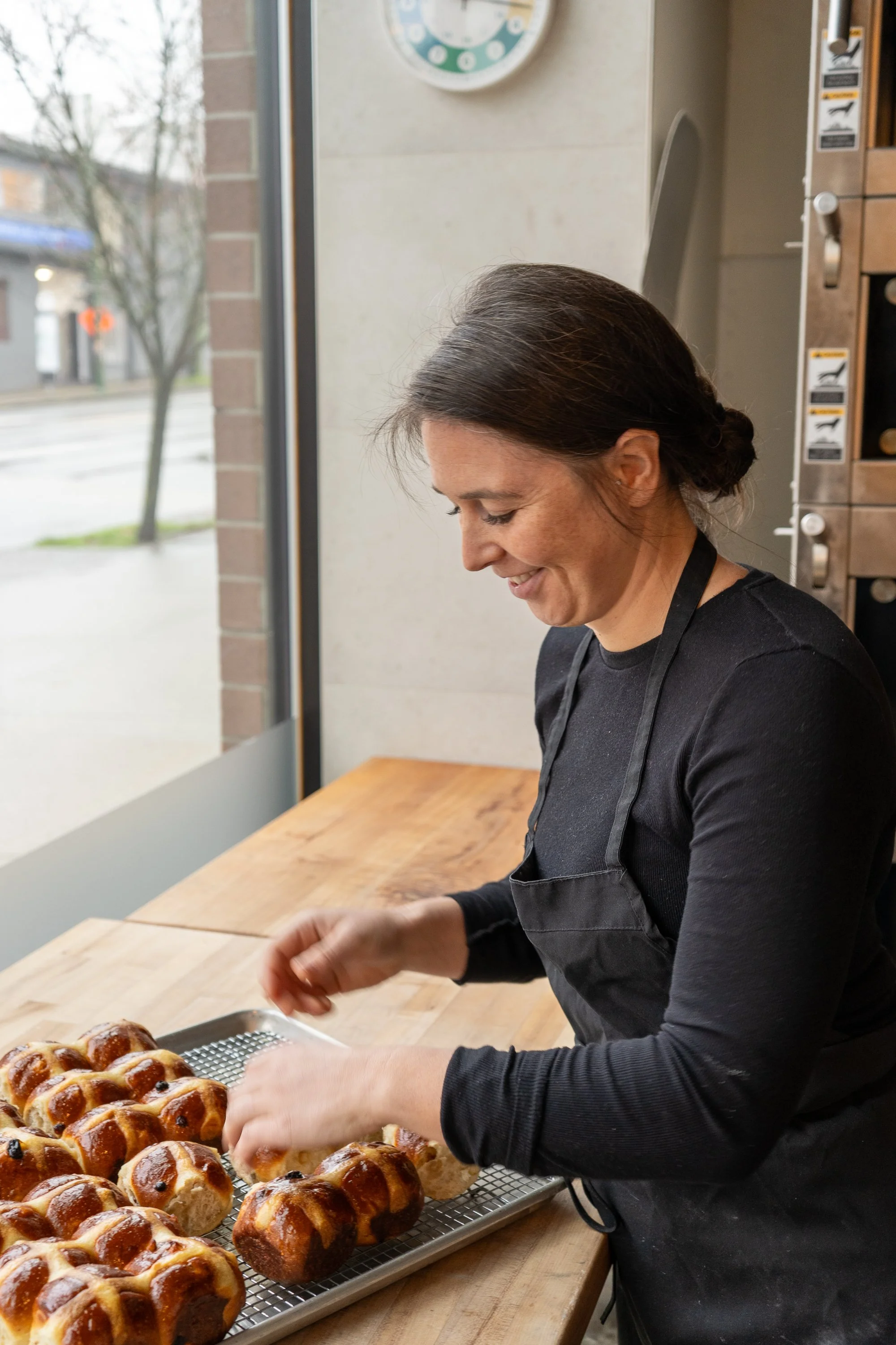 A woman in a black apron is preparing freshly baked bread or pastries on a wire rack inside a bakery or kitchen, with a large window to the outside in the background.