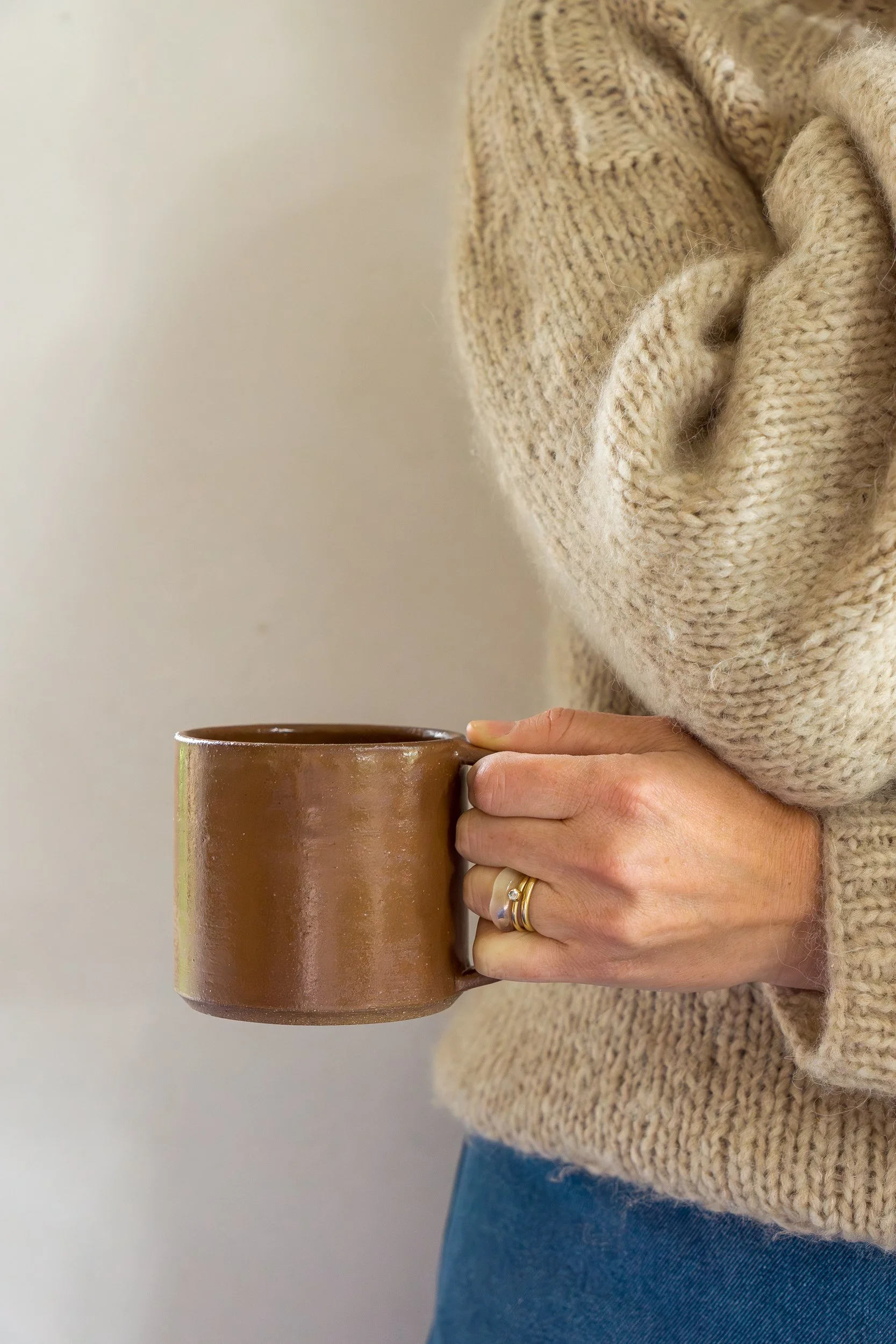 A person wearing a beige knit sweater holding a brown ceramic mug.