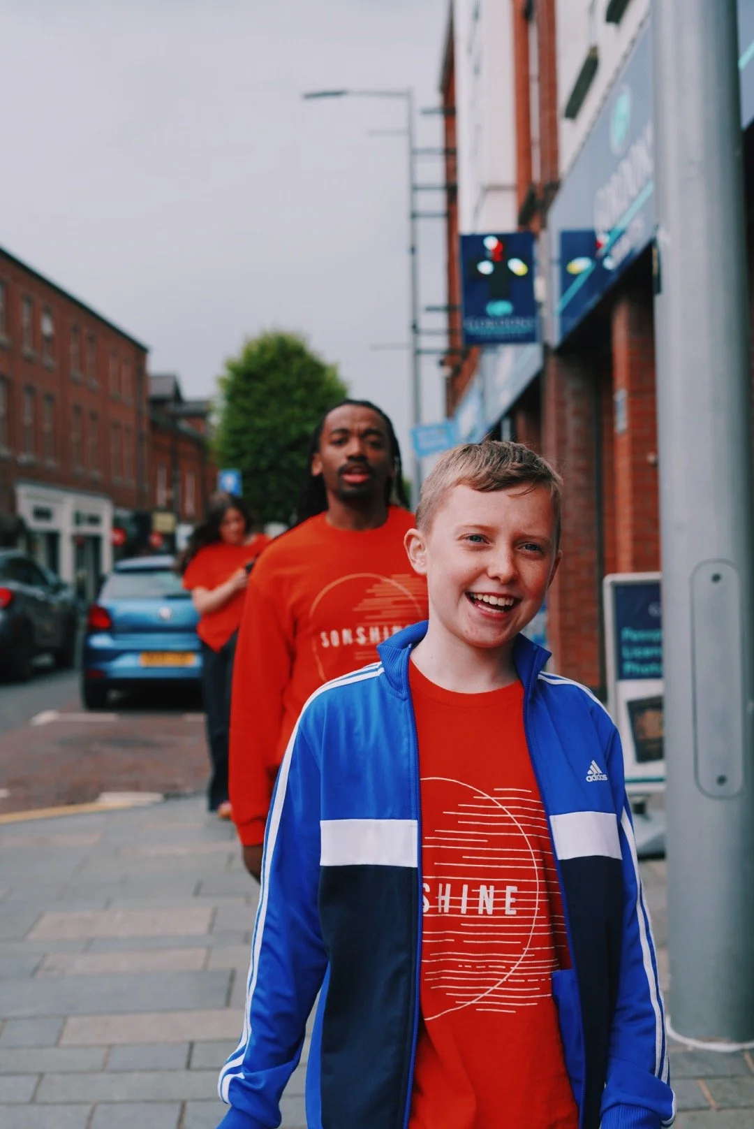 A group of young people in red t-shirts and casual jackets standing in a line on a city sidewalk, smiling and looking at the camera, with brick buildings and cars in the background.