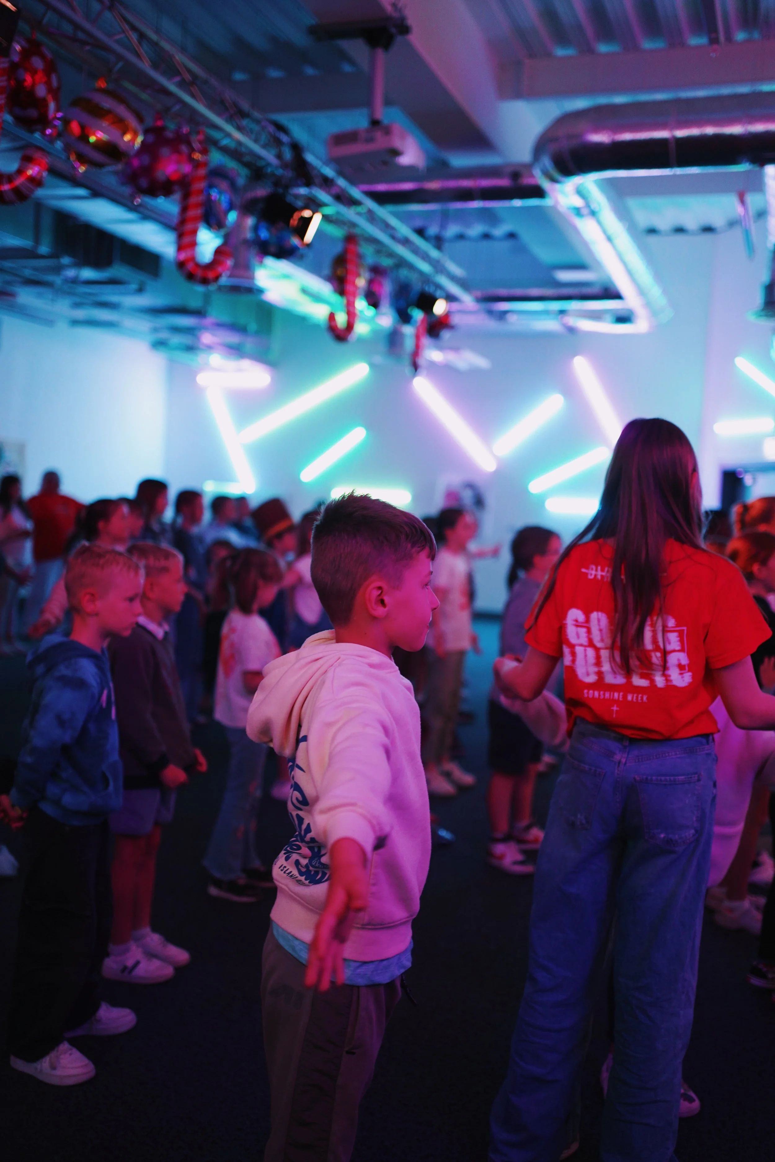 Children and adults standing in a dimly lit room with neon lights, likely at a dance or music event, facing towards a stage or performance area.