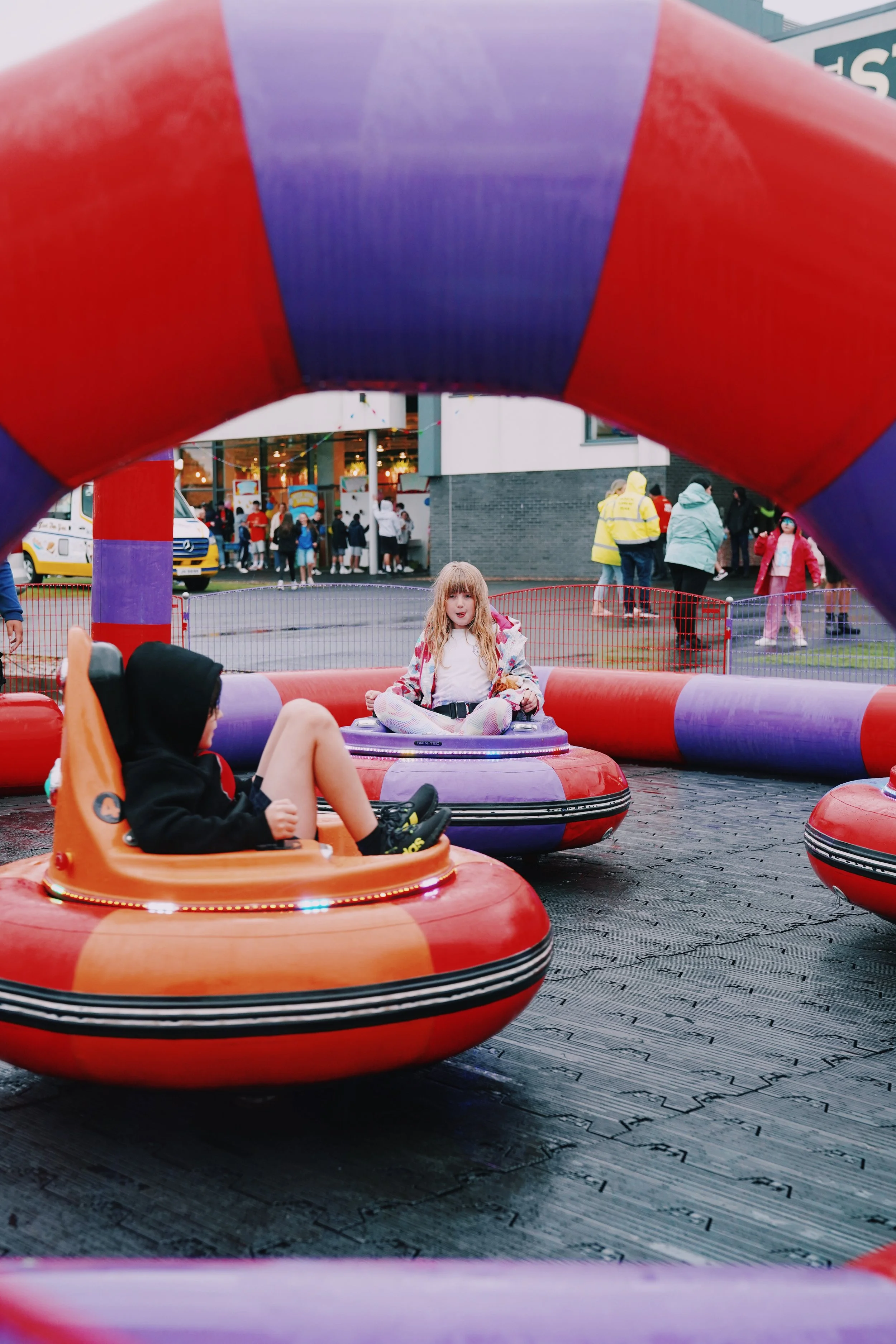 Children riding bumper cars at an amusement park, people walking in the background, and a colorful inflatable structure framing the scene.