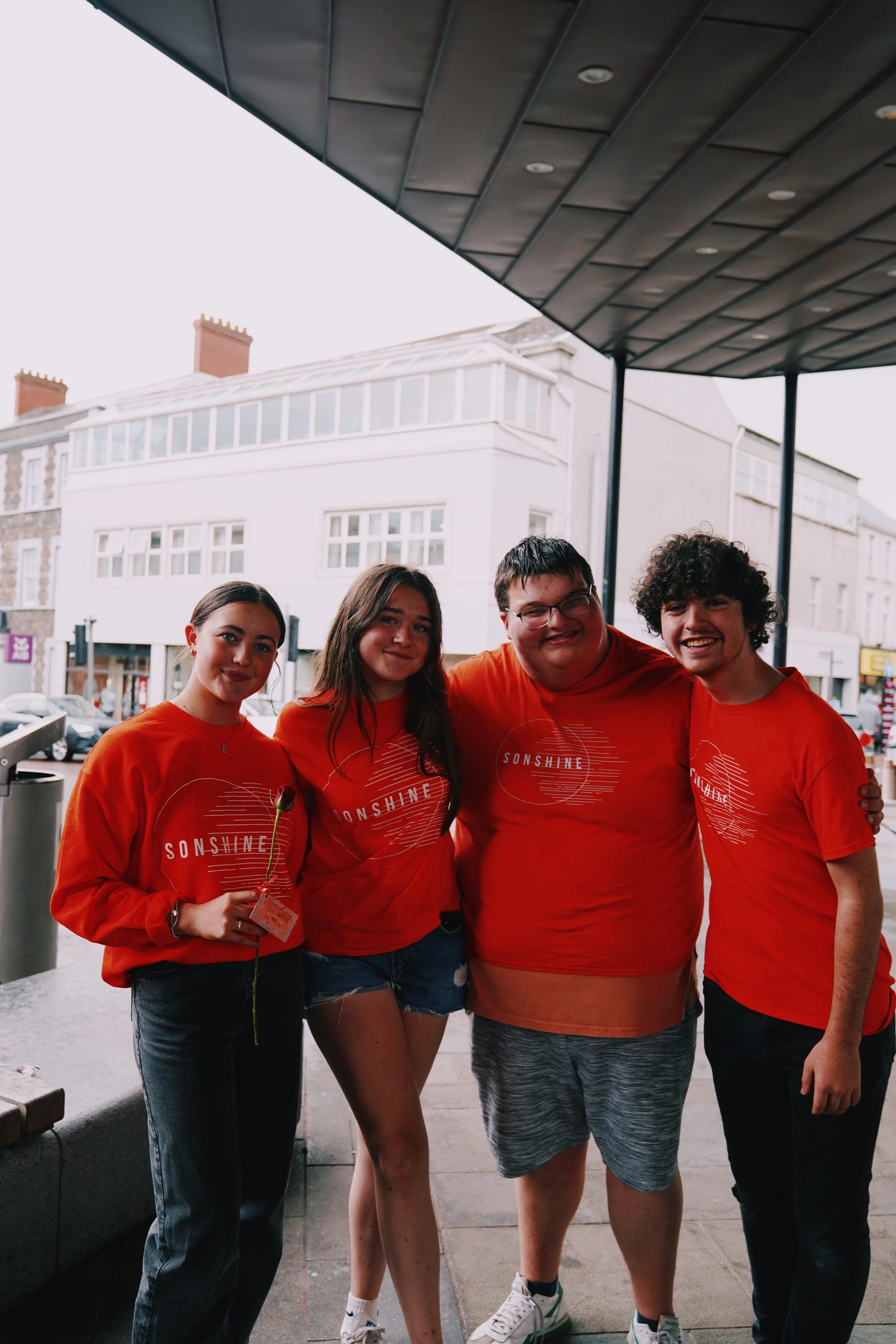 Group of five young people wearing matching red shirts with 'SUNSHINE' printed on them, standing outdoors on a city street, smiling for a photo.