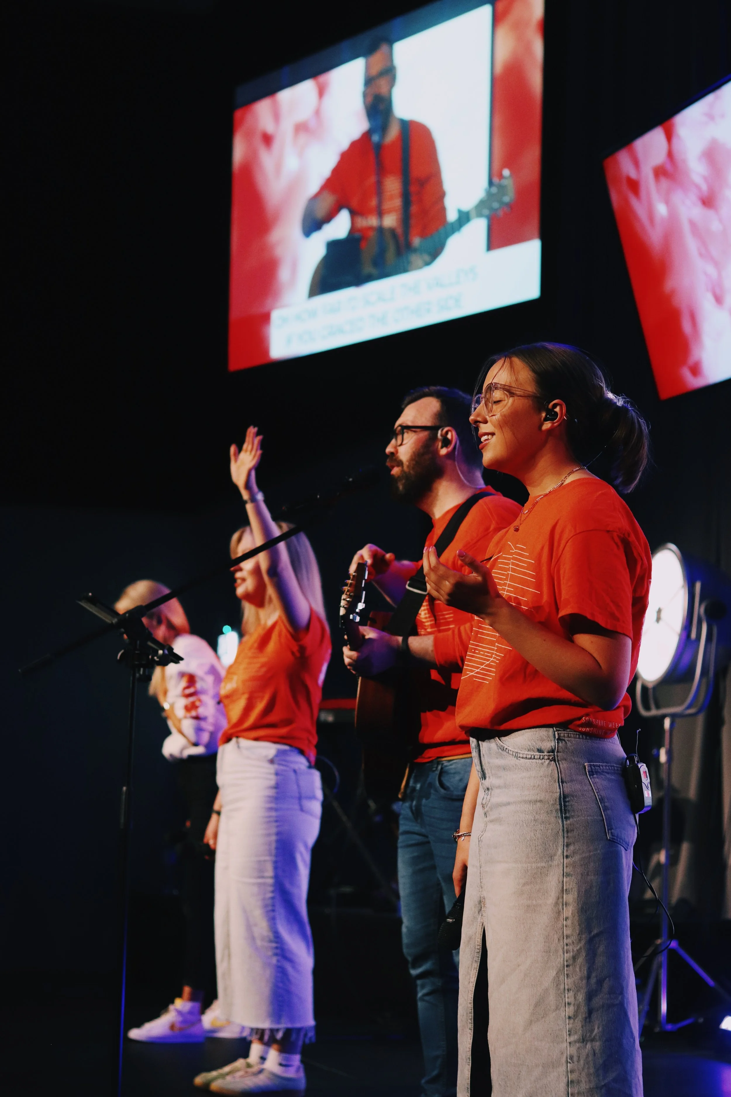 Group of people standing on stage, singing or praying with hands raised, dressed casually, illuminated by stage lights, with large screens showing a musician playing guitar in the background.