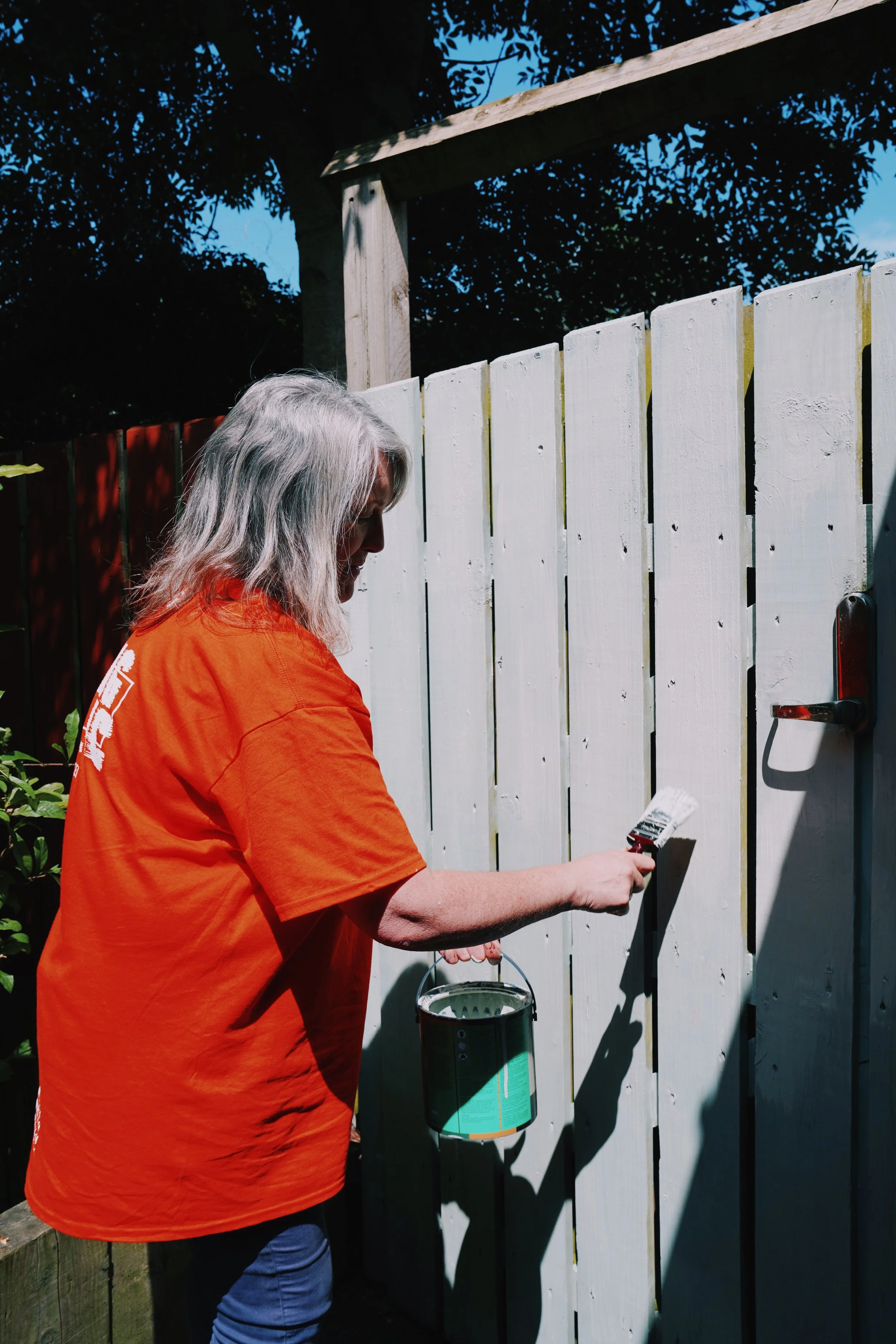 A person with long gray hair wearing an orange shirt painting a white wooden fence with a paintbrush under a blue sky.