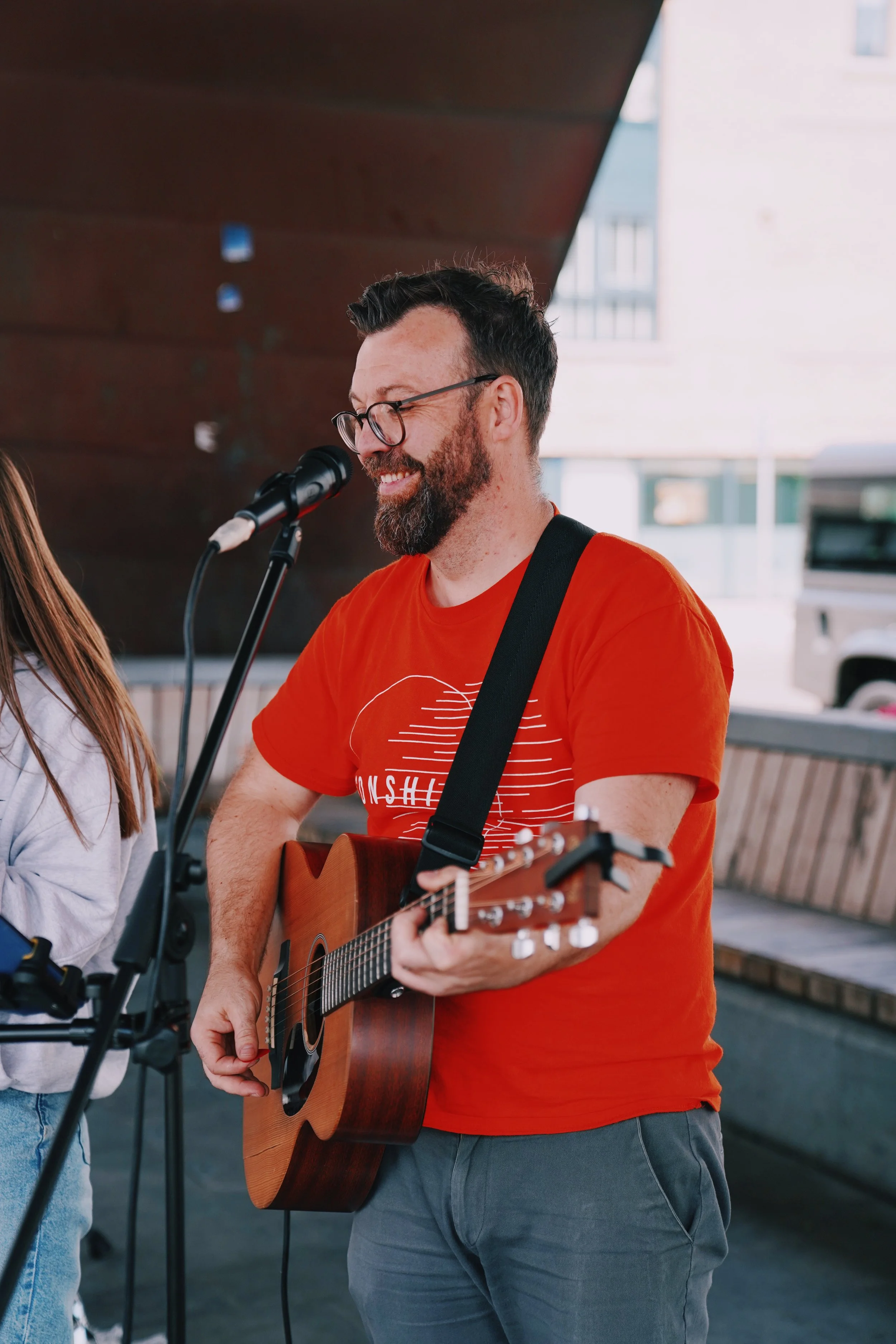 A man with glasses and a beard wearing an orange t-shirt is playing an acoustic guitar and singing into a microphone, outdoors near a building.