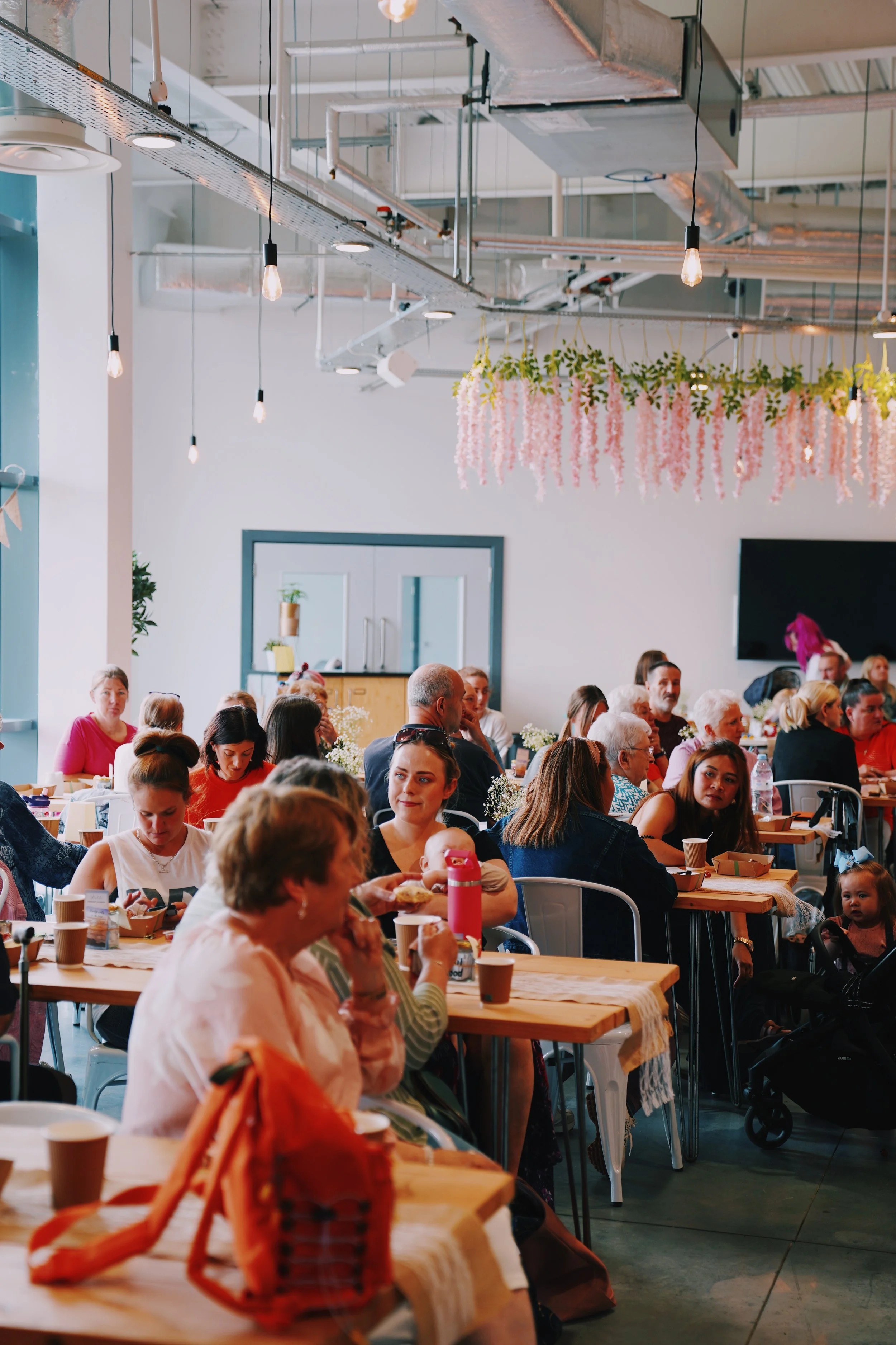 A large indoor gathering of diverse people, mostly women, seated at long wooden tables, sharing a meal in a decorated event space with hanging pendant lights and pink floral decorations.