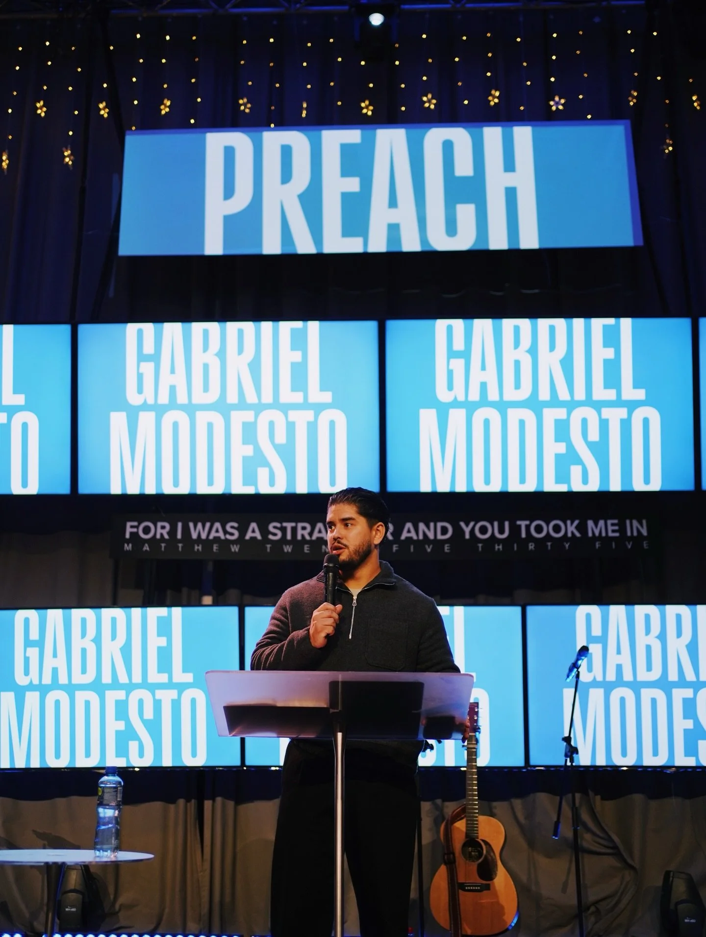 A man speaking into a microphone at a stage with a music stand, guitar, and water bottle. Behind him is a large screen displaying the words 'GABRIEL MODOSTO' and a smaller screen with the word 'PREACH'. The background features dark curtains with small yellow flower decorations.