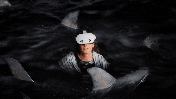 Person wearing virtual reality goggles surrounded by shark fins in dark water.