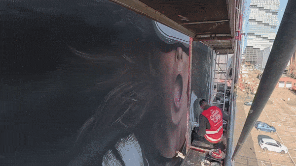 A person working on a large mural on the side of a building, depicting a close-up of a person's face with an open mouth. The worker is sitting on a small platform with tools nearby, city buildings and parked cars visible in the background.