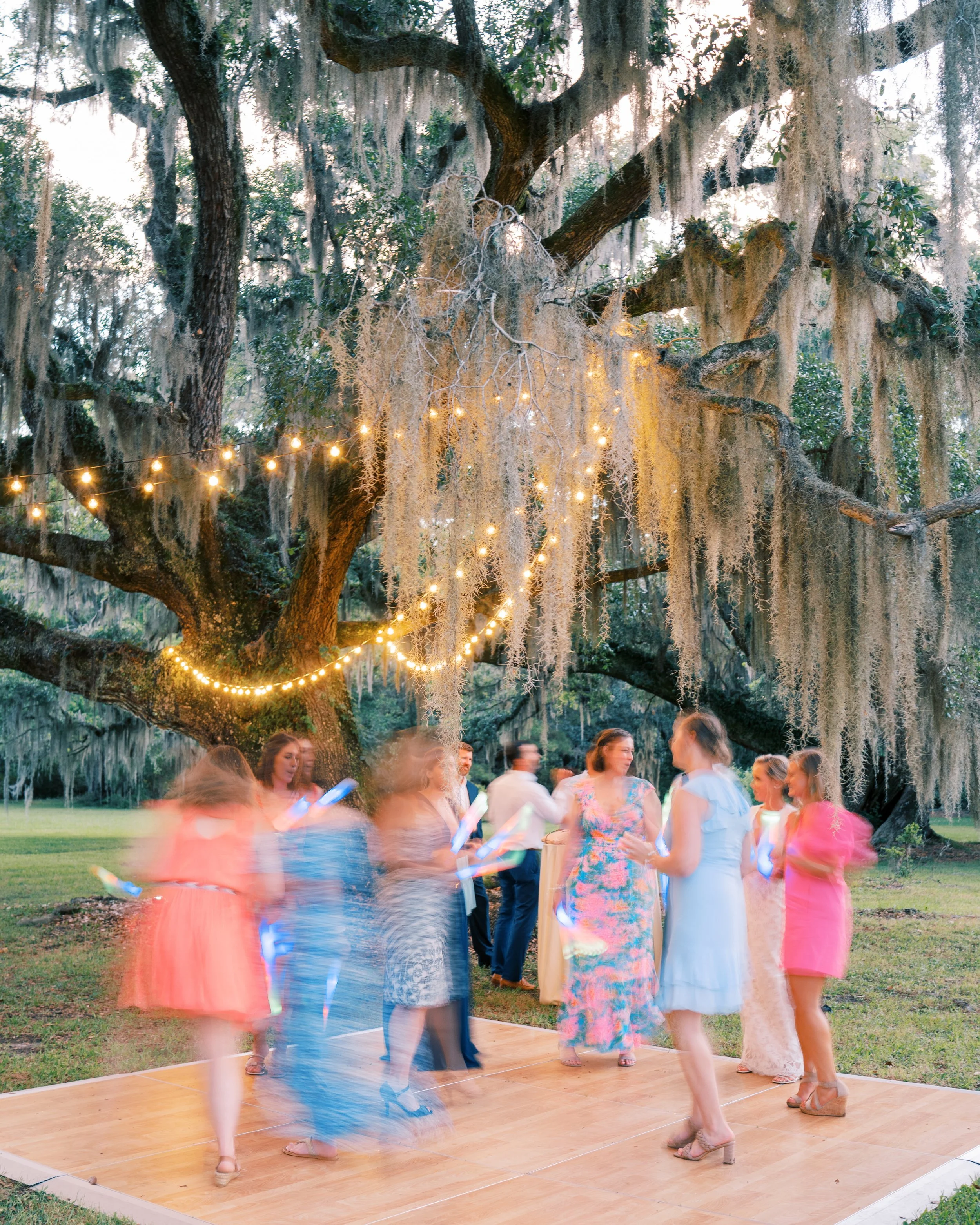 People dancing outdoors under string lights and Spanish moss on a wooden platform.