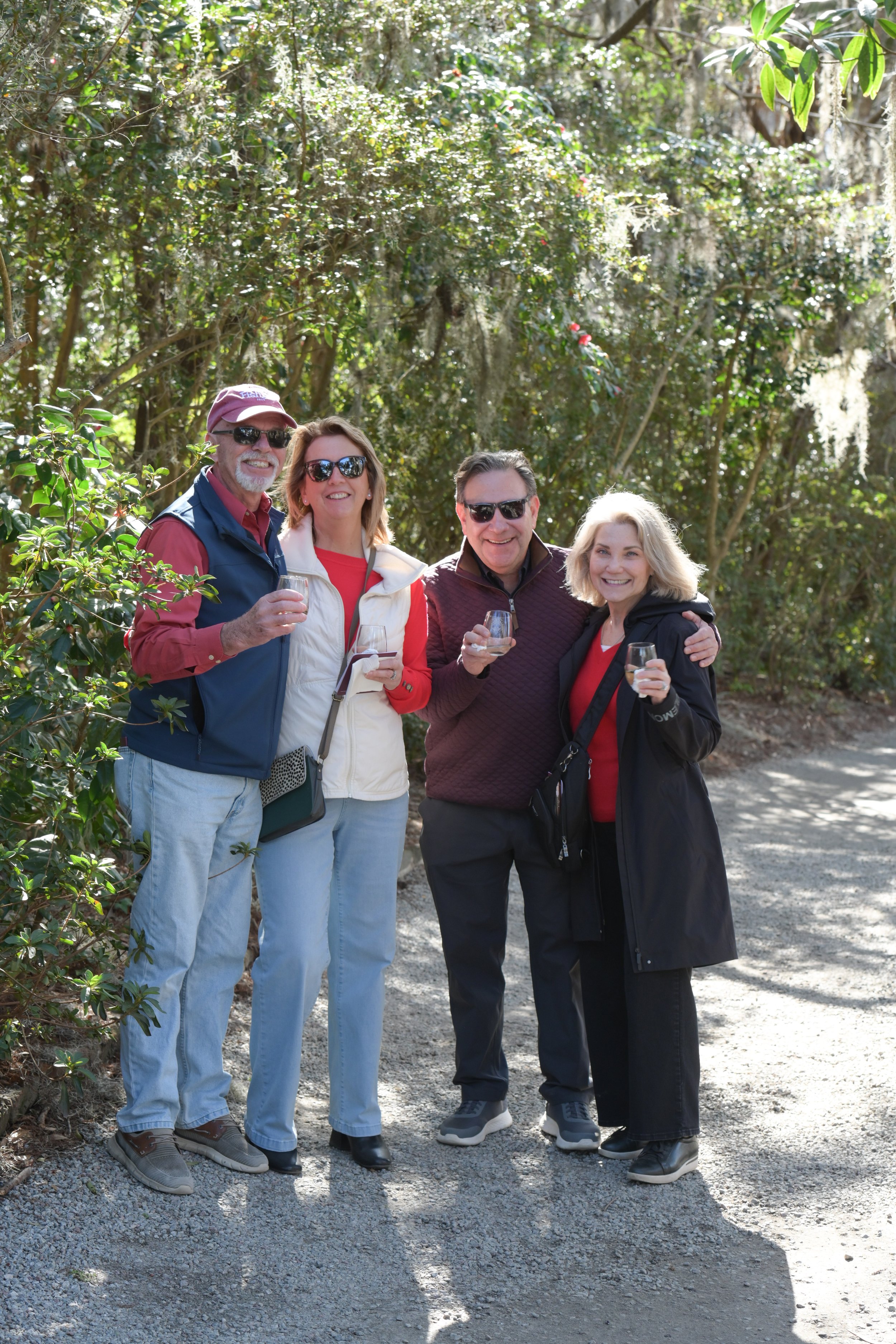 Two smiling couples stand in line along a pathway surrounded by trees.  They are each holding a glass of wine from the International Wine and Sweets Stroll.