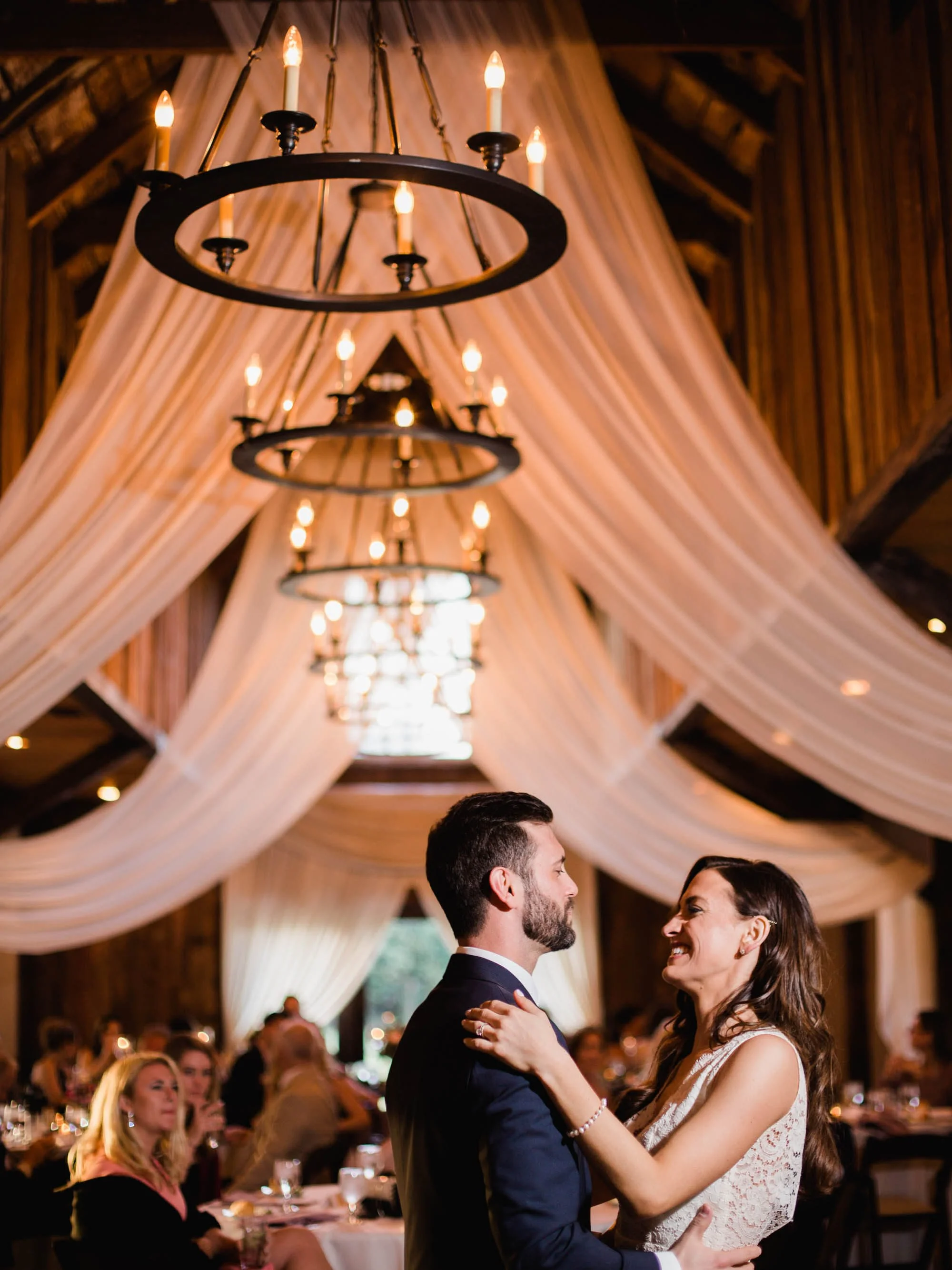 A couple dancing at a wedding reception in a rustic venue with draped fabric on the ceiling and chandeliers. Guests seated at tables in the background.