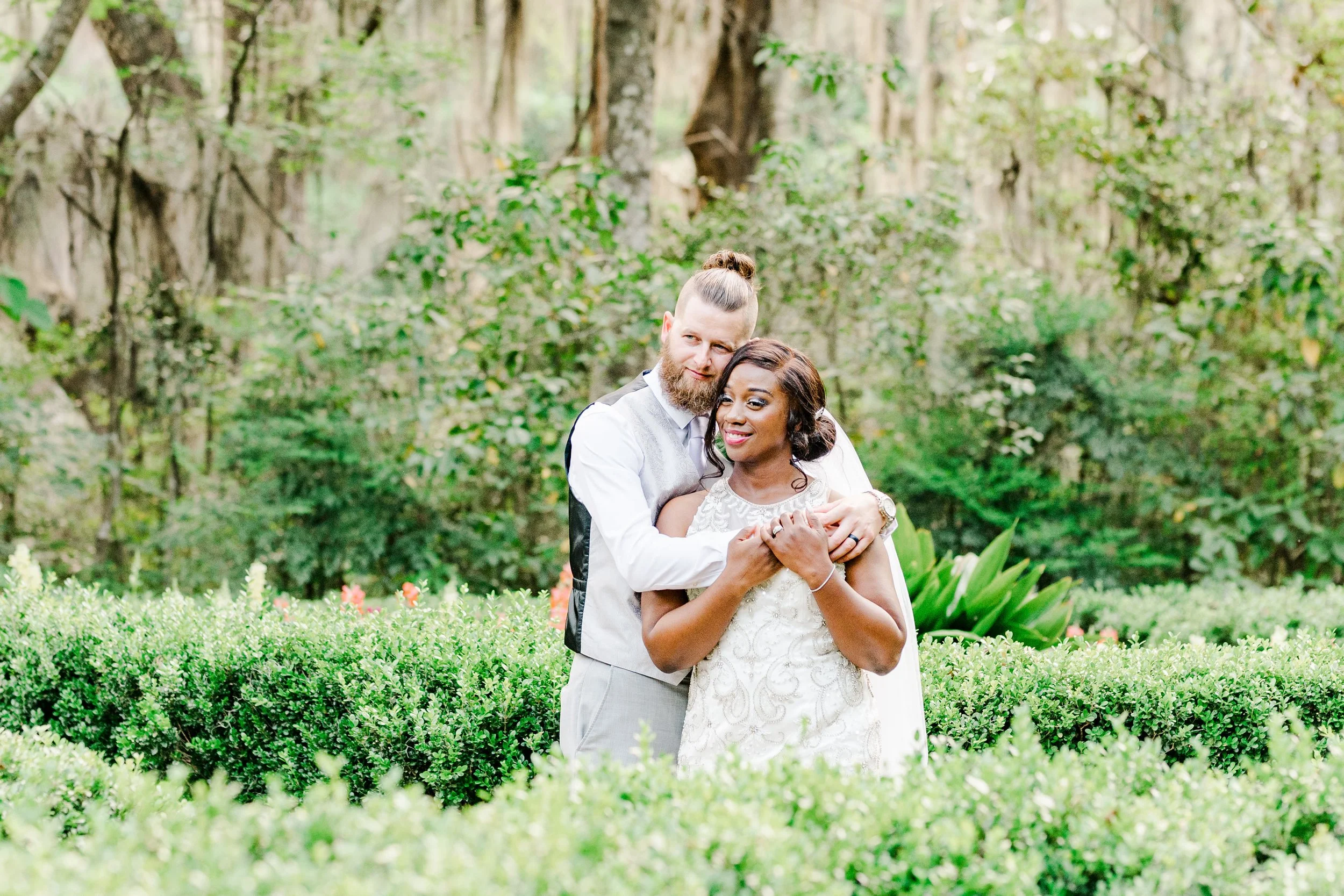 Bride and groom embracing in a garden setting with green foliage and trees in the background.