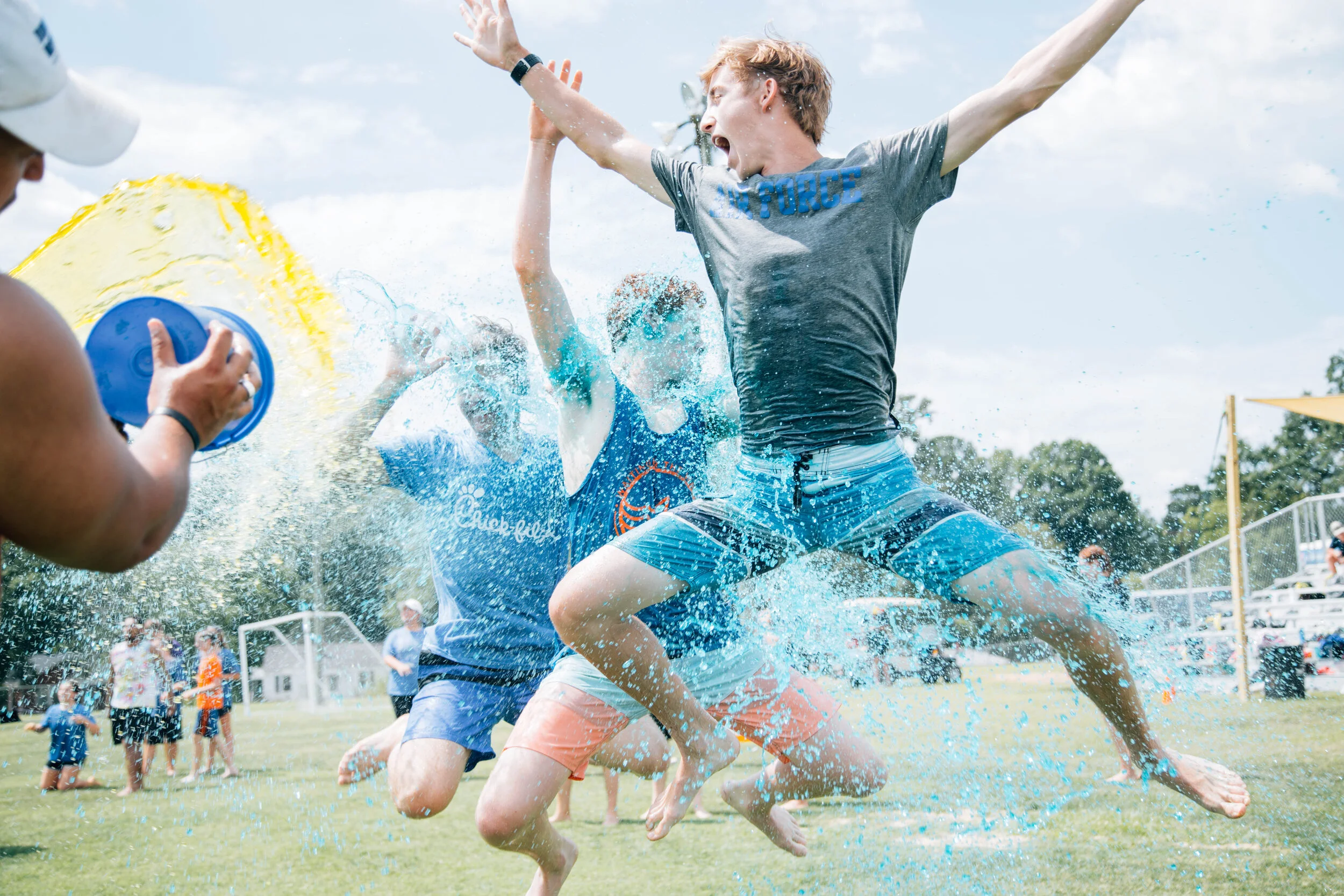 People participating in a water fight during a summer event at a park, with a person in a grey t-shirt and blue shorts jumping and splashing water, others in the background holding water toys and playing.