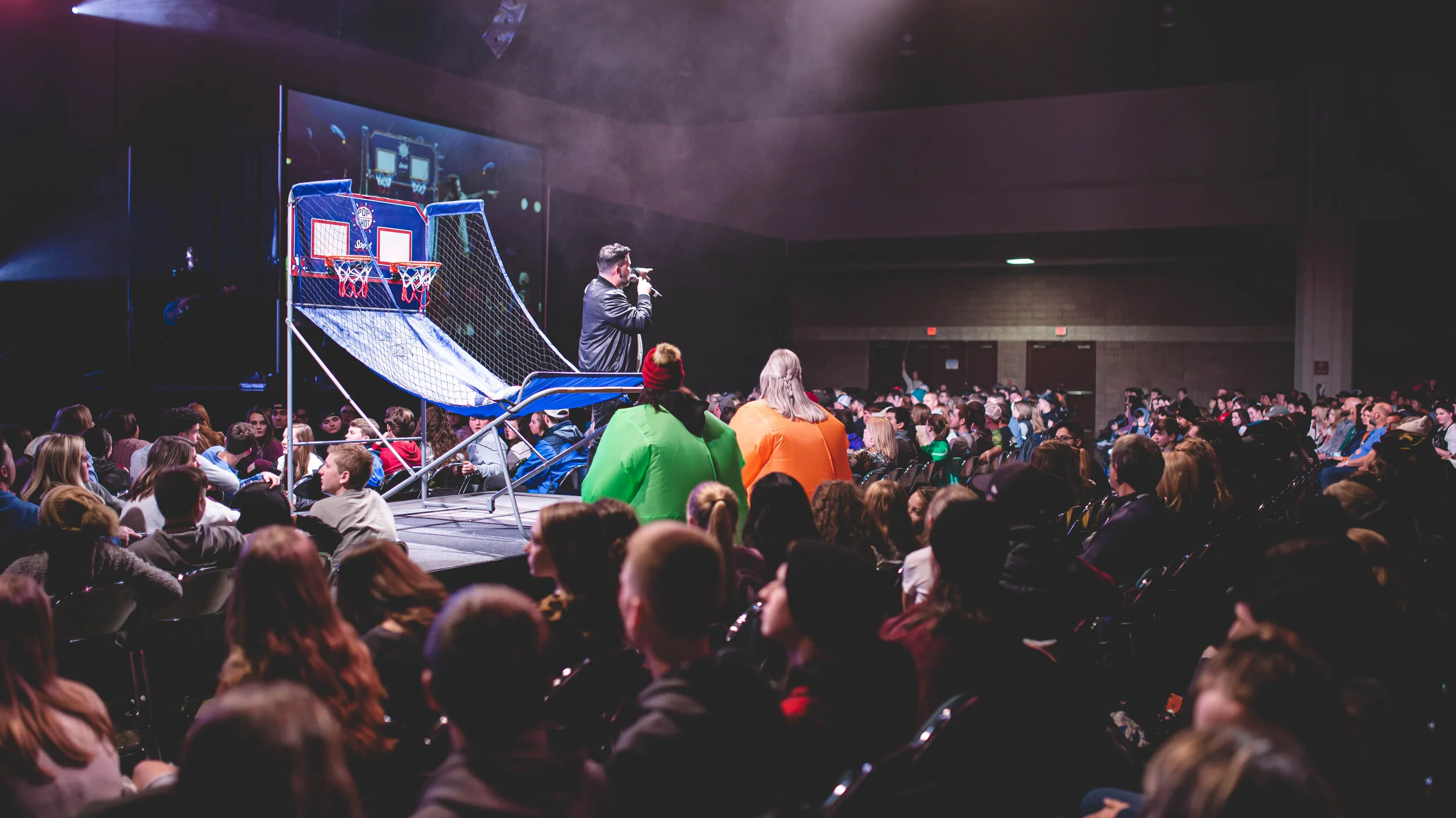 An indoor basketball arcade game setup on a stage with a large crowd seated in rows watching. A speaker in a leather jacket is addressing the audience, and there are colorful tents in the audience.