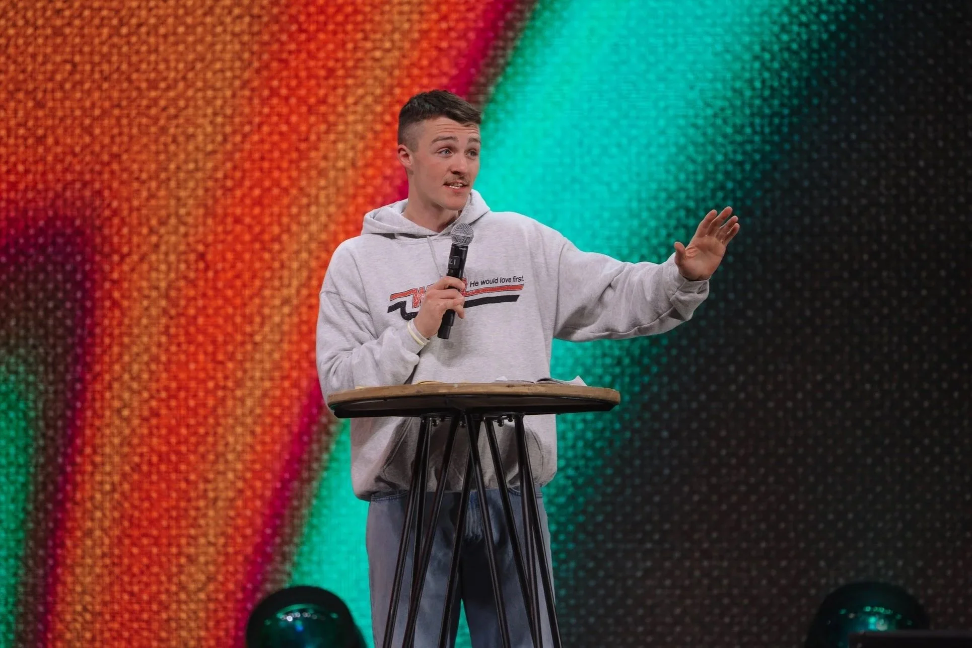 A young man wearing a gray hoodie with text on it, holding a microphone, and speaking while standing behind a round wooden table on a stage with a colorful, abstract background.