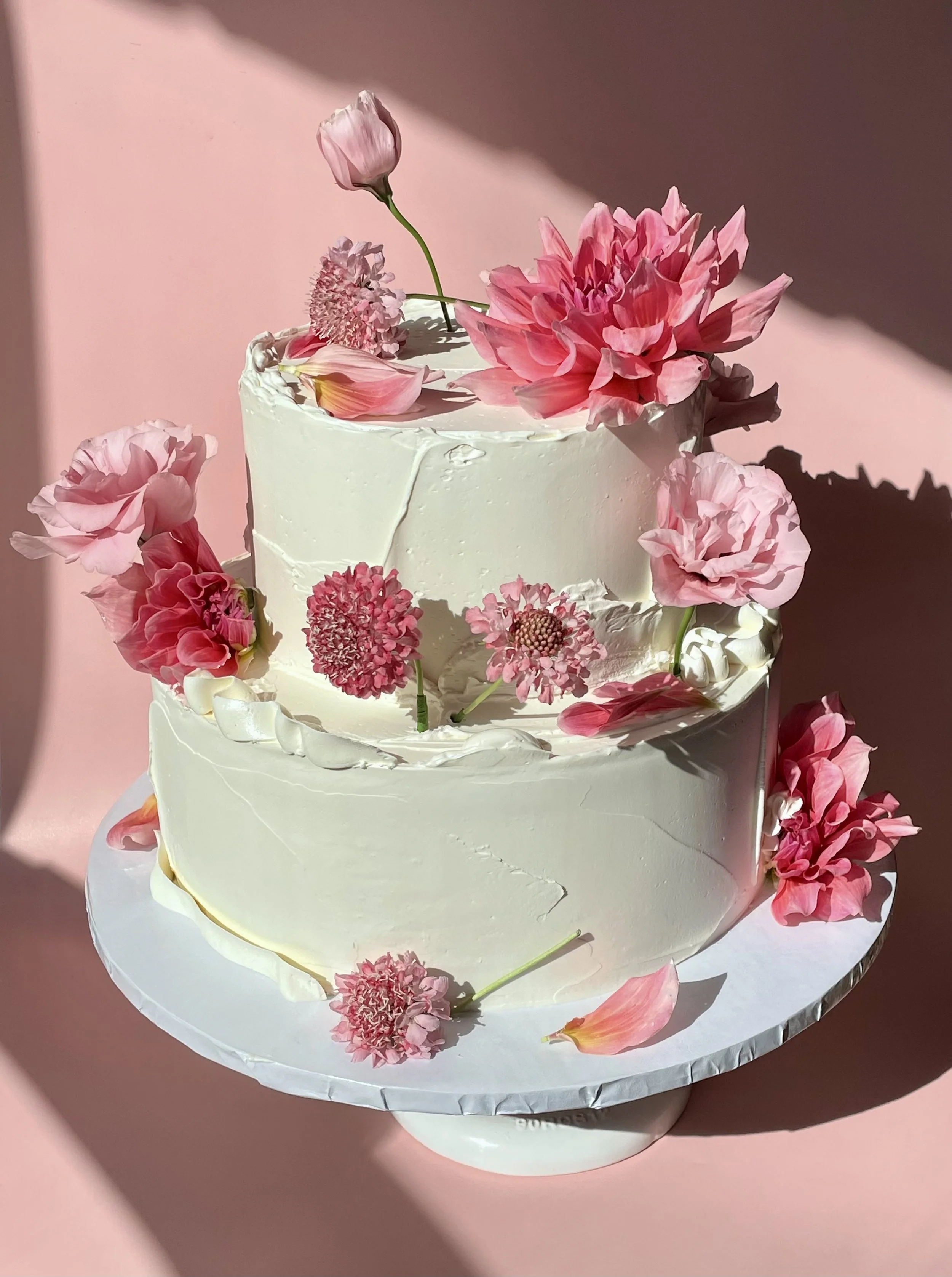 A white, two-tiered cake decorated with pink flowers and petals against a pink background.