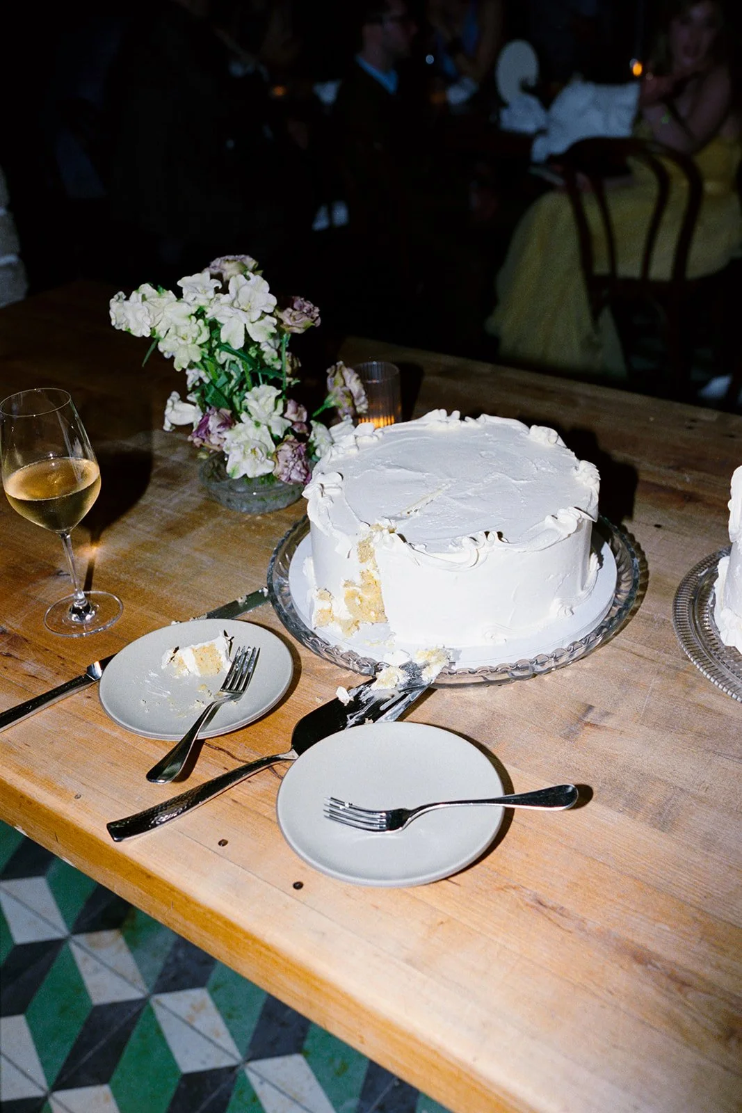 A partially eaten white cake on a glass cake stand with a slice taken out, surrounded by empty plates, forks, a knife, a glass of white wine, and a flower arrangement in a vase on a wooden table, in a dimly lit setting with people in the background.