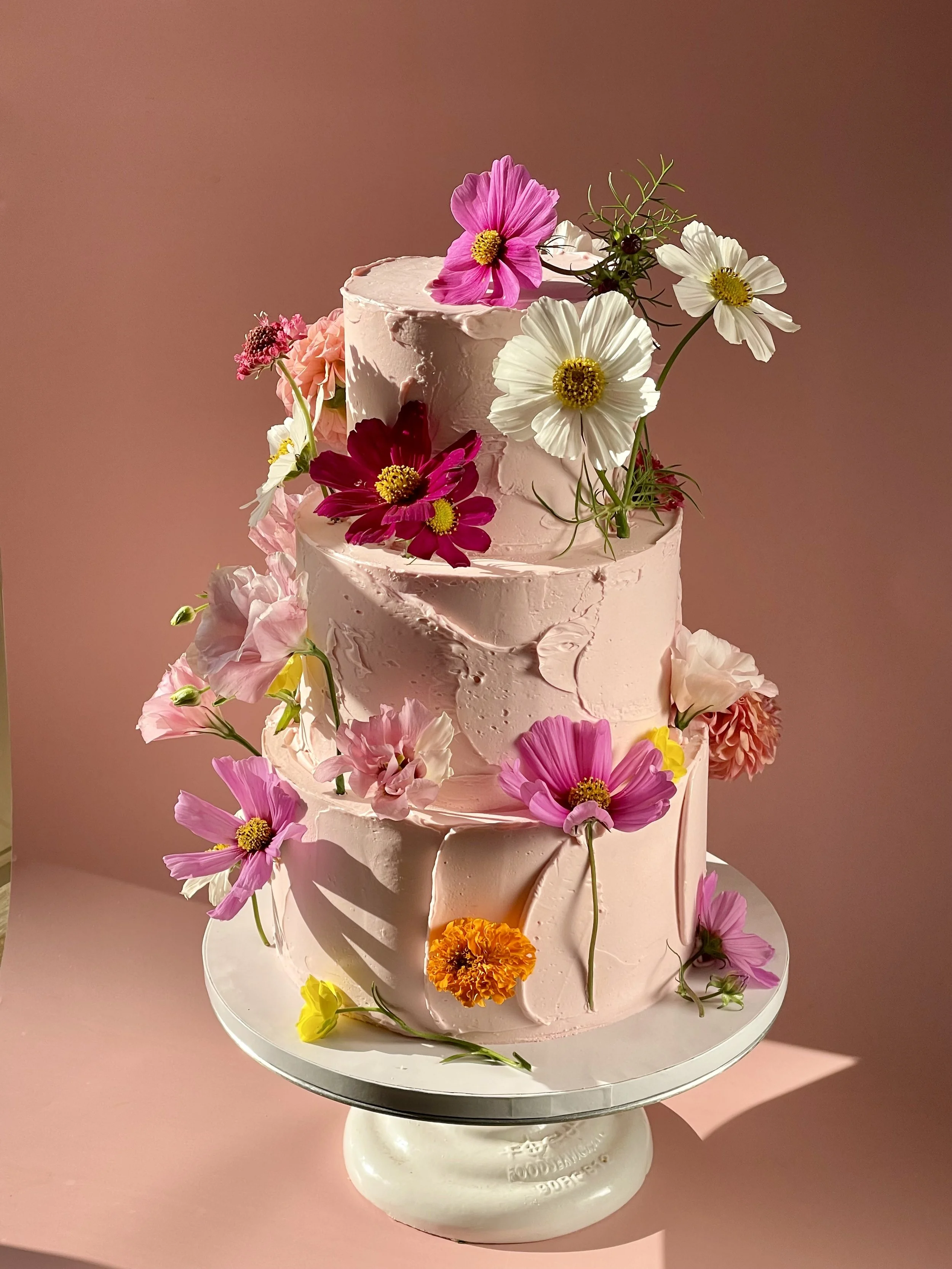 A pink, three-tiered cake decorated with various colorful flowers including pink, white, and orange blossoms, set against a pink background.