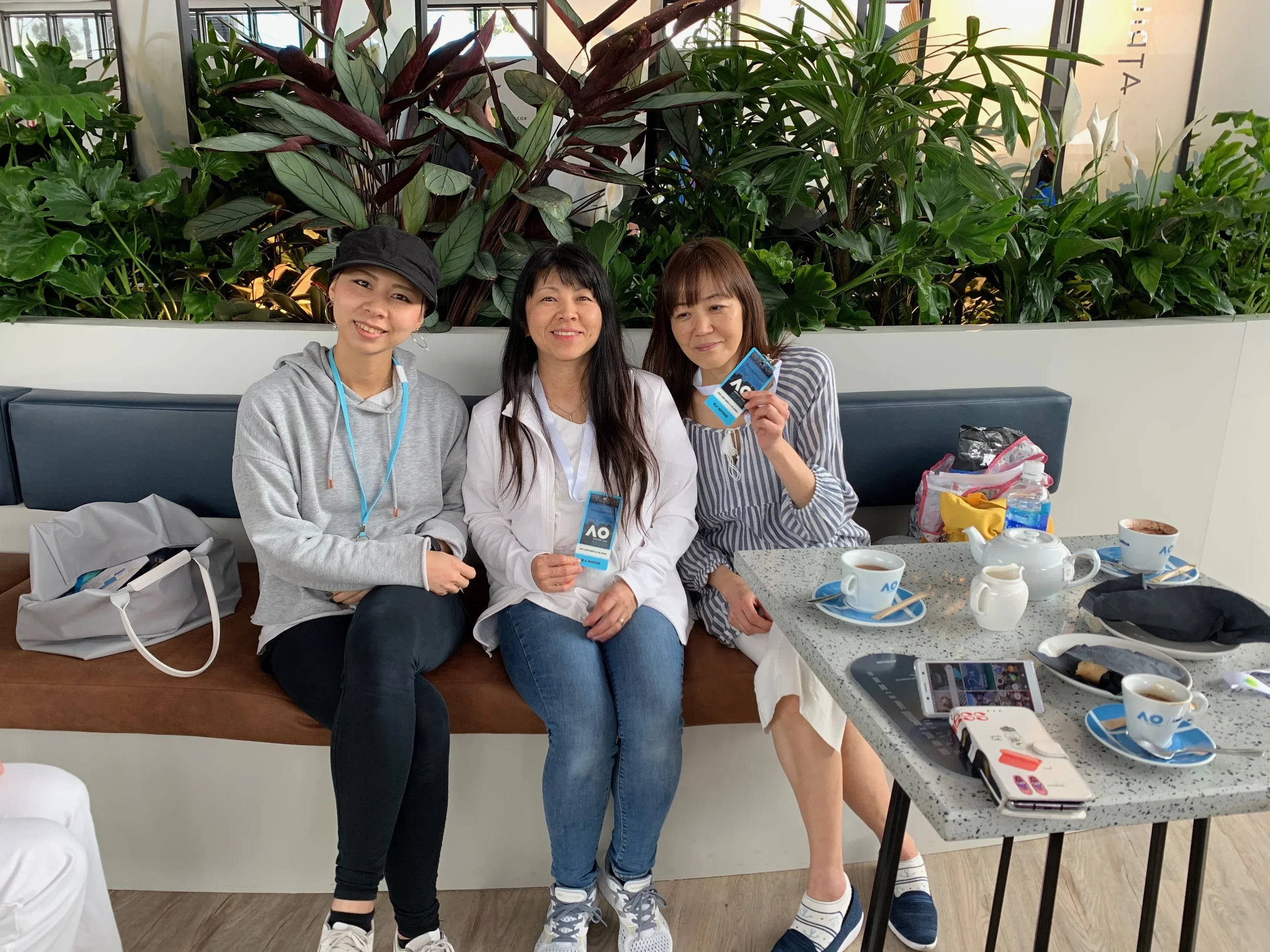 Three women sitting on a brown bench in a cafe with a background of green plants, a table with tea cups, a teapot, and various items, with two women holding AO cards and smiling at the camera.