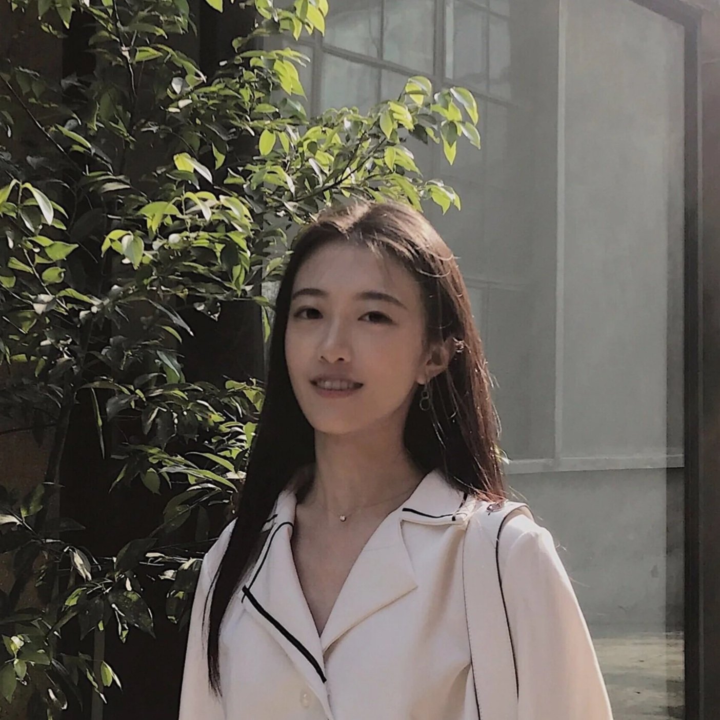 A young woman with long dark hair smiling outdoors near a green leafy bush and glass building, wearing a cream-colored shirt with dark piping.