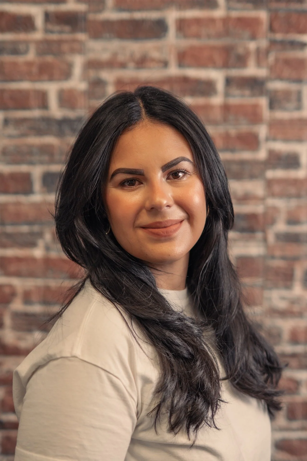 A woman with long dark hair and a beige top smiling in front of a brick wall.