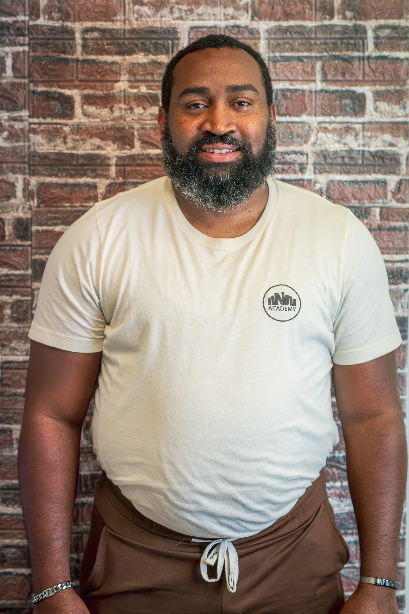 A man with a beard and short hair standing against a brick wall, wearing a black t-shirt with a logo on the chest.