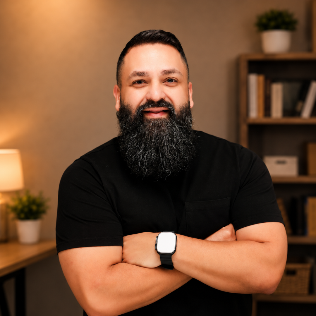 A man with a full beard and short dark hair, wearing a black shirt and a smartwatch, standing with arms crossed against a dark background.