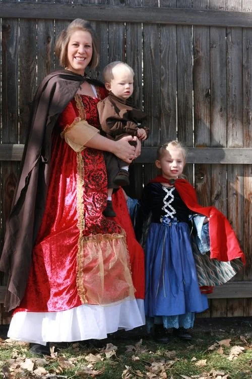 A woman and two children dressed in costumes for a medieval or fantasy-themed event, standing outdoors in front of a wooden fence.