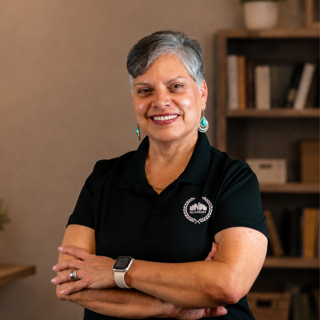 A middle-aged woman with short gray hair and hoop earrings, smiling, wearing a black t-shirt with a logo that says 'NDA Academy,' standing in front of a brick wall.