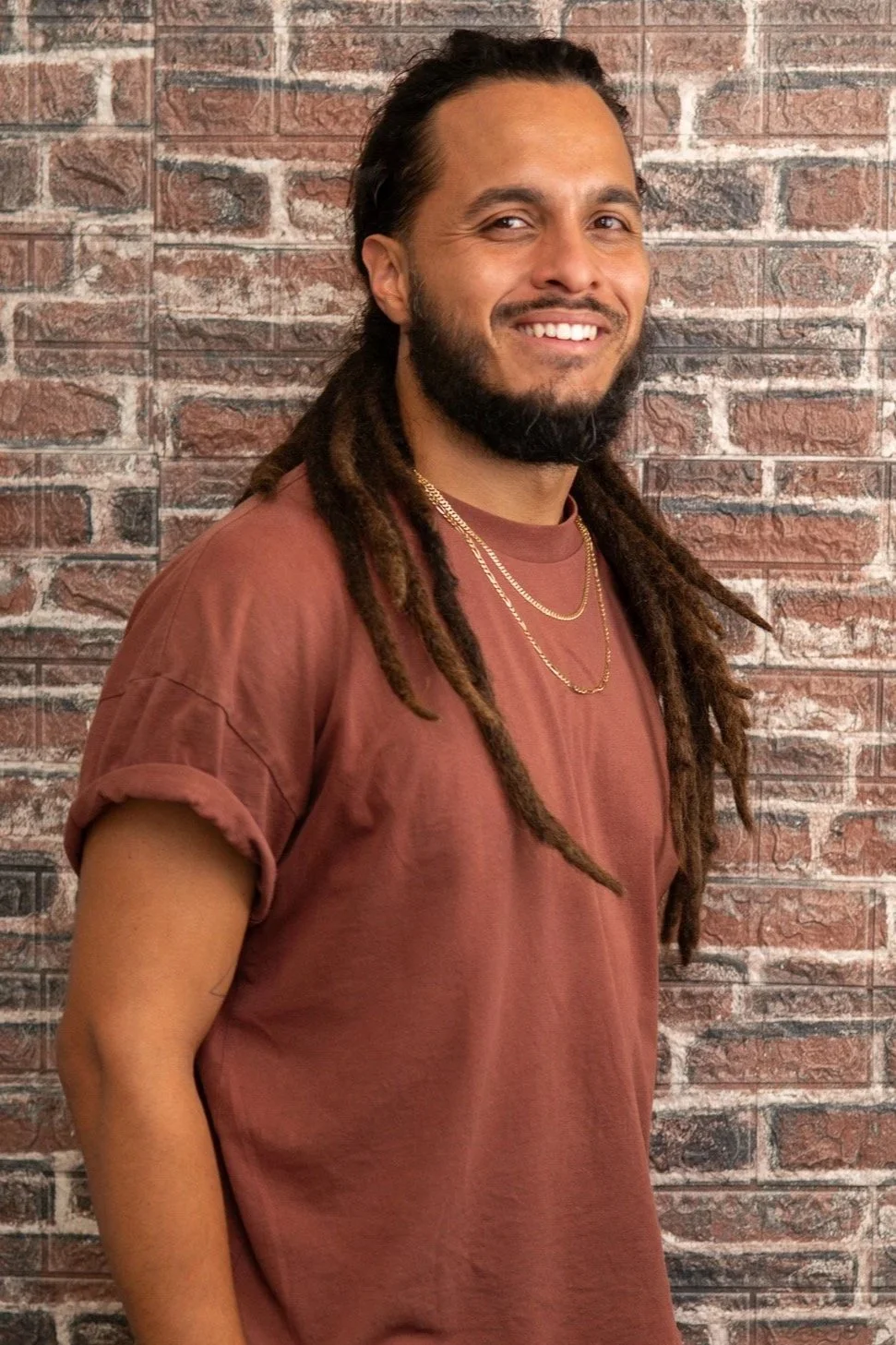 A man with long dreadlocks, beard, and mustache smiling in front of a brick wall, wearing a green Teenage Mutant Ninja Turtles T-shirt.