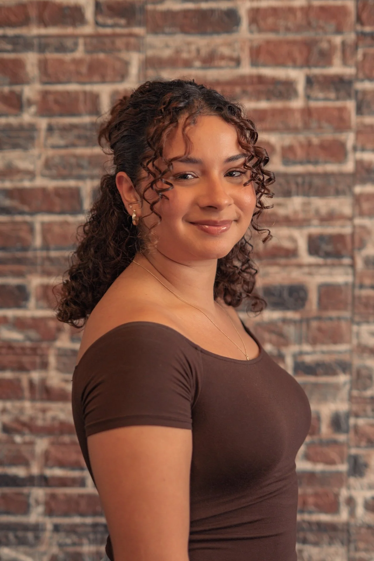 A woman with curly brown hair and light brown skin, smiling, wearing a brown top and jewelry, standing in front of a brick wall.