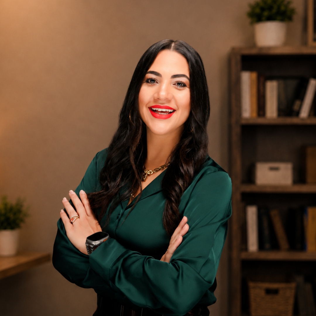 A smiling woman with long dark wavy hair, wearing a green satin blouse, gold jewelry, a smartwatch, and gold hoop earrings, standing against a black background.