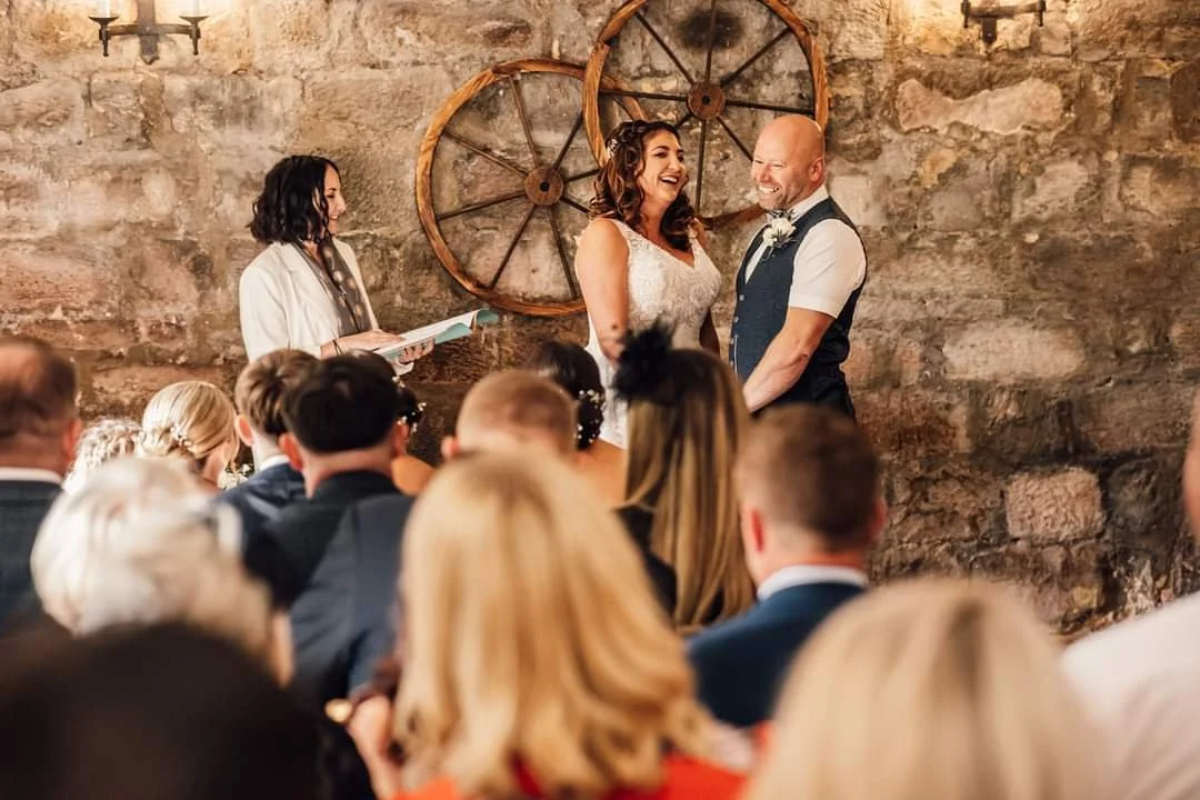 A couple gets married in front of friends and family in a rustic venue with stone walls and wooden wagon wheels, including Joanne McLaughlin, The Celebrant of the North, award-winning north east celebrant.