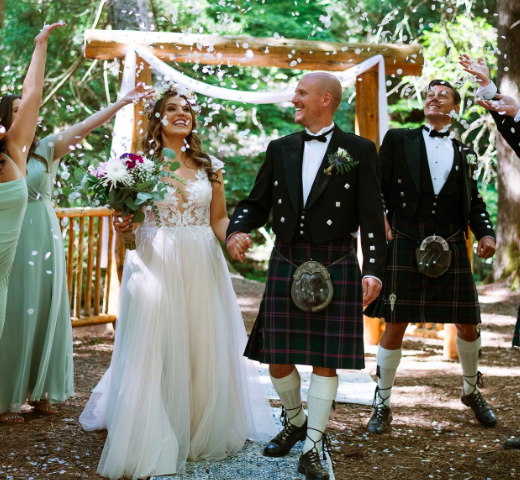 Wedding celebration in Ganaraska Forest, Ontario, Canada, officiated by Joanne McLaughlin, The Celebrant of the North. Award winning North East Celebrant. Bride and groom walking hand in hand outdoors with guests celebrating, throwing flower petals.