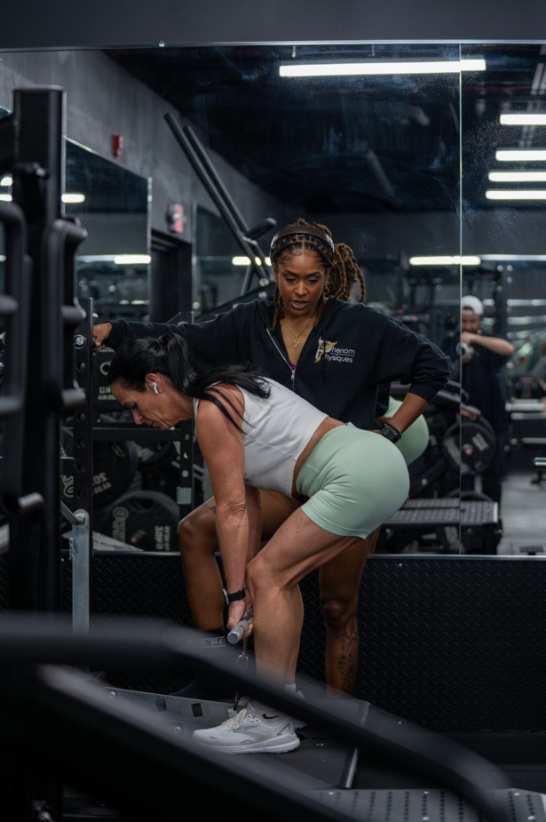 A personal trainer assisting a woman with a workout at the gym, with weightlifting equipment and mirrors in the background.