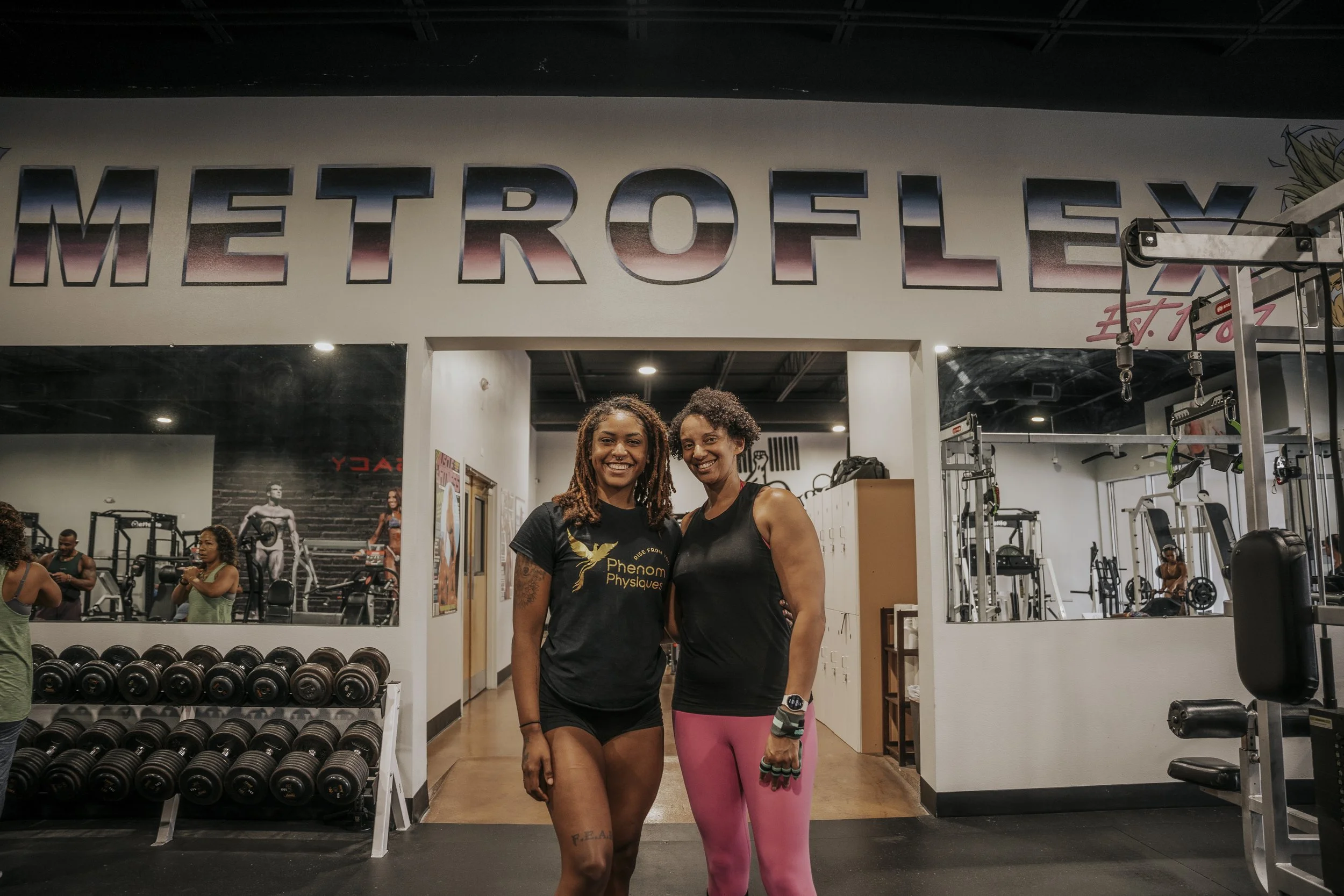 Two women standing together inside a gym in front of a large sign that says 'METROFLEX'. One woman is wearing a black t-shirt with a logo, and the other is in a black tank top and pink leggings. gym equipment, weights, and other gym-goers are visible