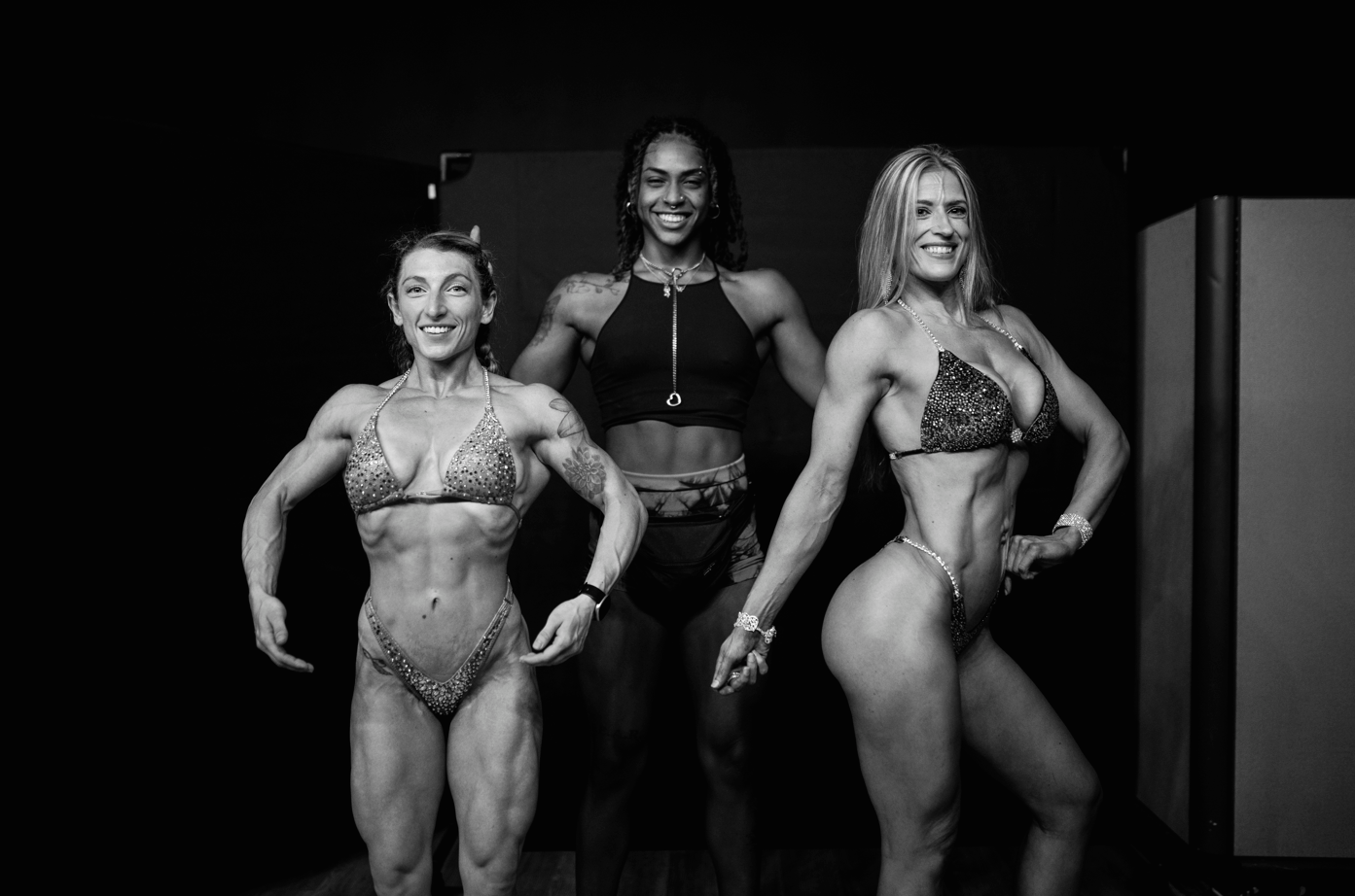 Three women in bikinis and athletic outfits posing and smiling in a bodybuilding competition setting.