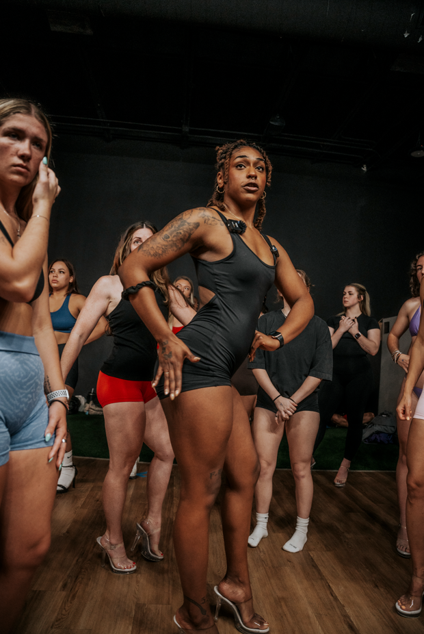 A group of women gathered on a wooden floor in a dark indoor space, with one woman in the center wearing a black dress and high heels, posing with her hands on her hips, surrounded by other women in casual and athletic attire.