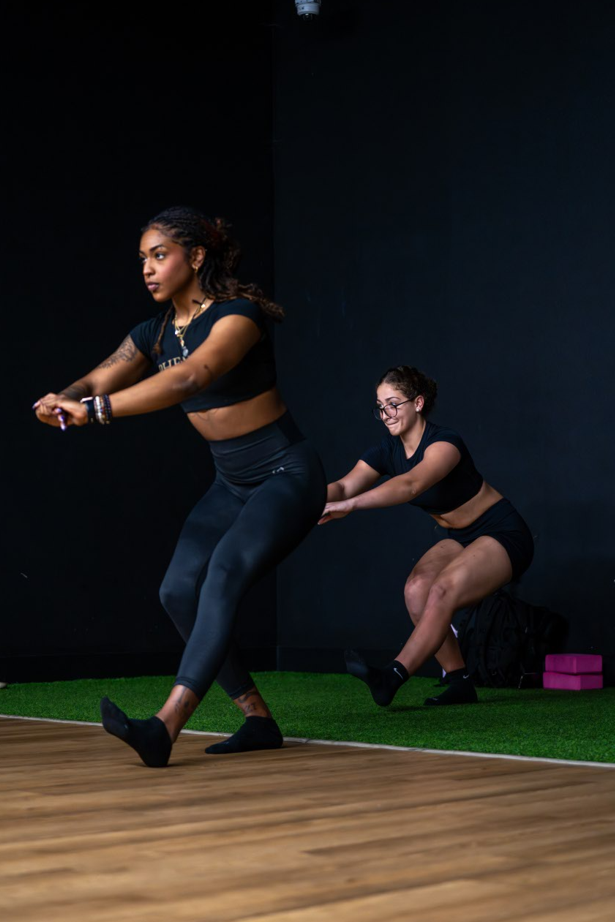 Two women in black workout clothes pulling on a resistance band in a gym, with one woman in front and the other kneeling behind her, holding the band.