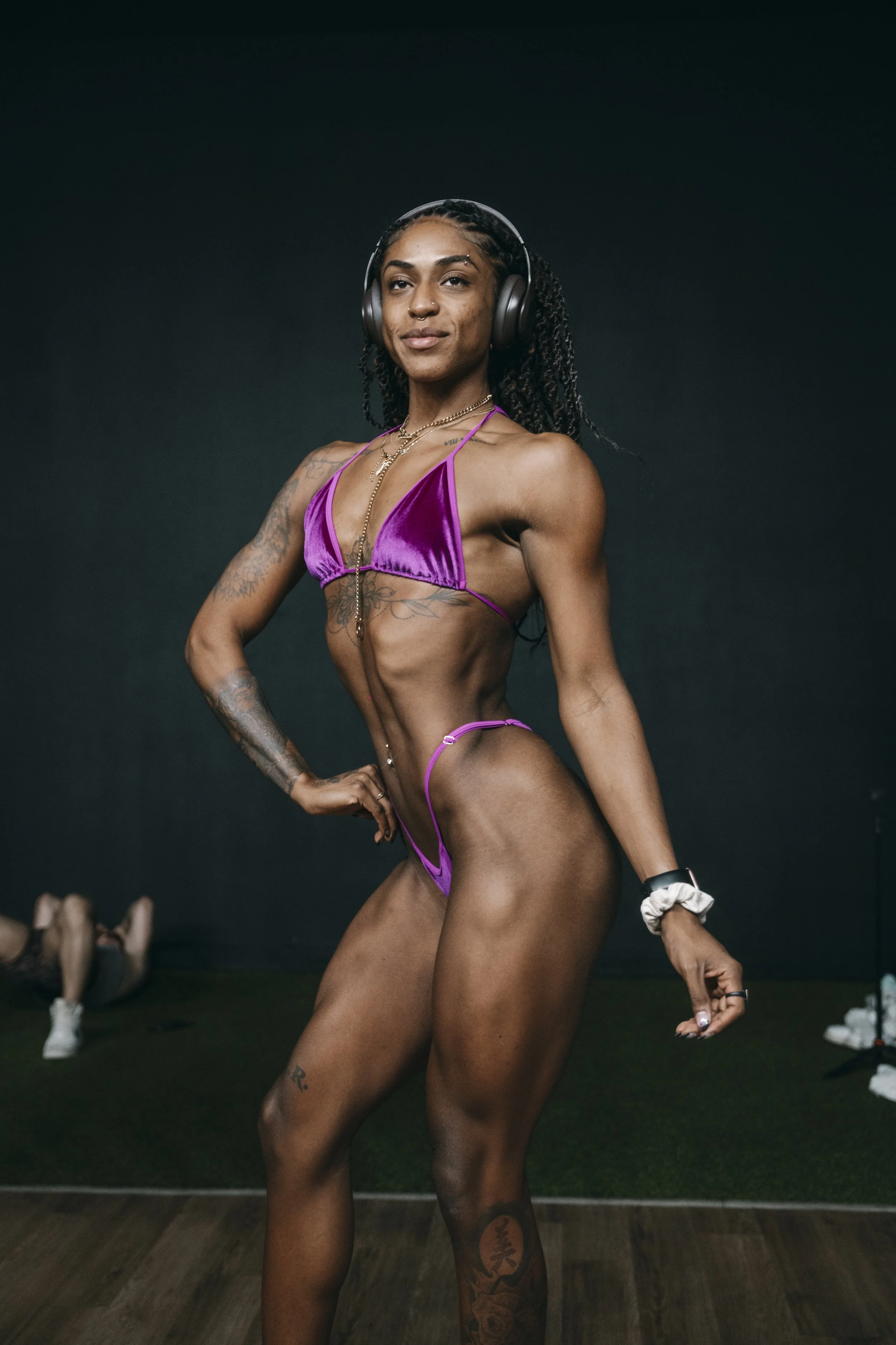 A fit woman with tan skin and tattoos on her left arm and right thigh wearing purple athletic swimwear, headphones, and jewelry, standing in front of a dark background.