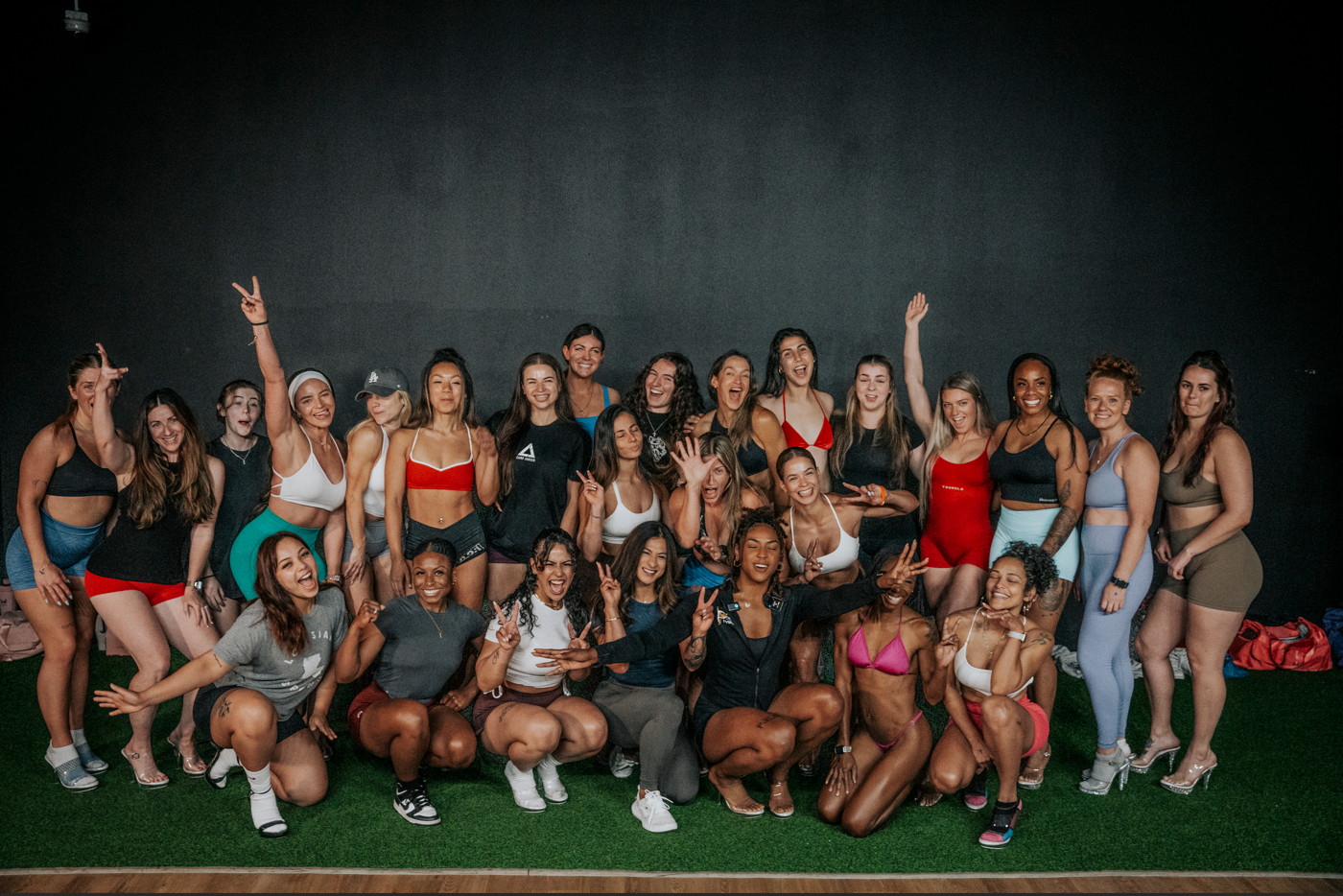 Group of women in athletic wear posing joyfully for a photo on a green indoor turf in front of a dark background.