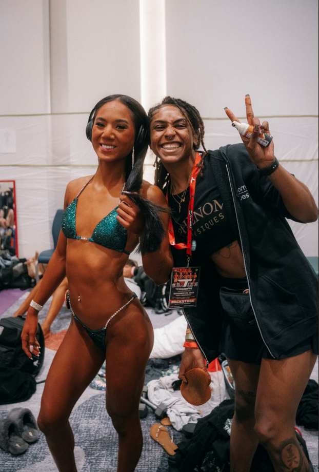 A woman in a sparkly teal bikini posing backstage at a fitness competition, smiling, with a woman beside her making a peace sign and holding a spray bottle.