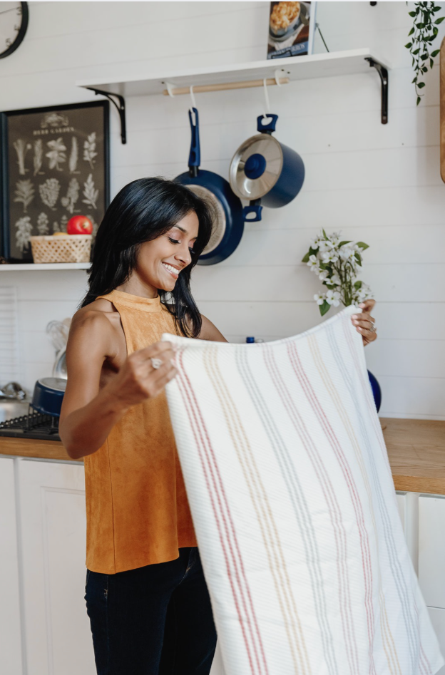 Woman smiling and folding a cloth in a kitchen with hanging pots, a white shelf, and a framed botanical print.