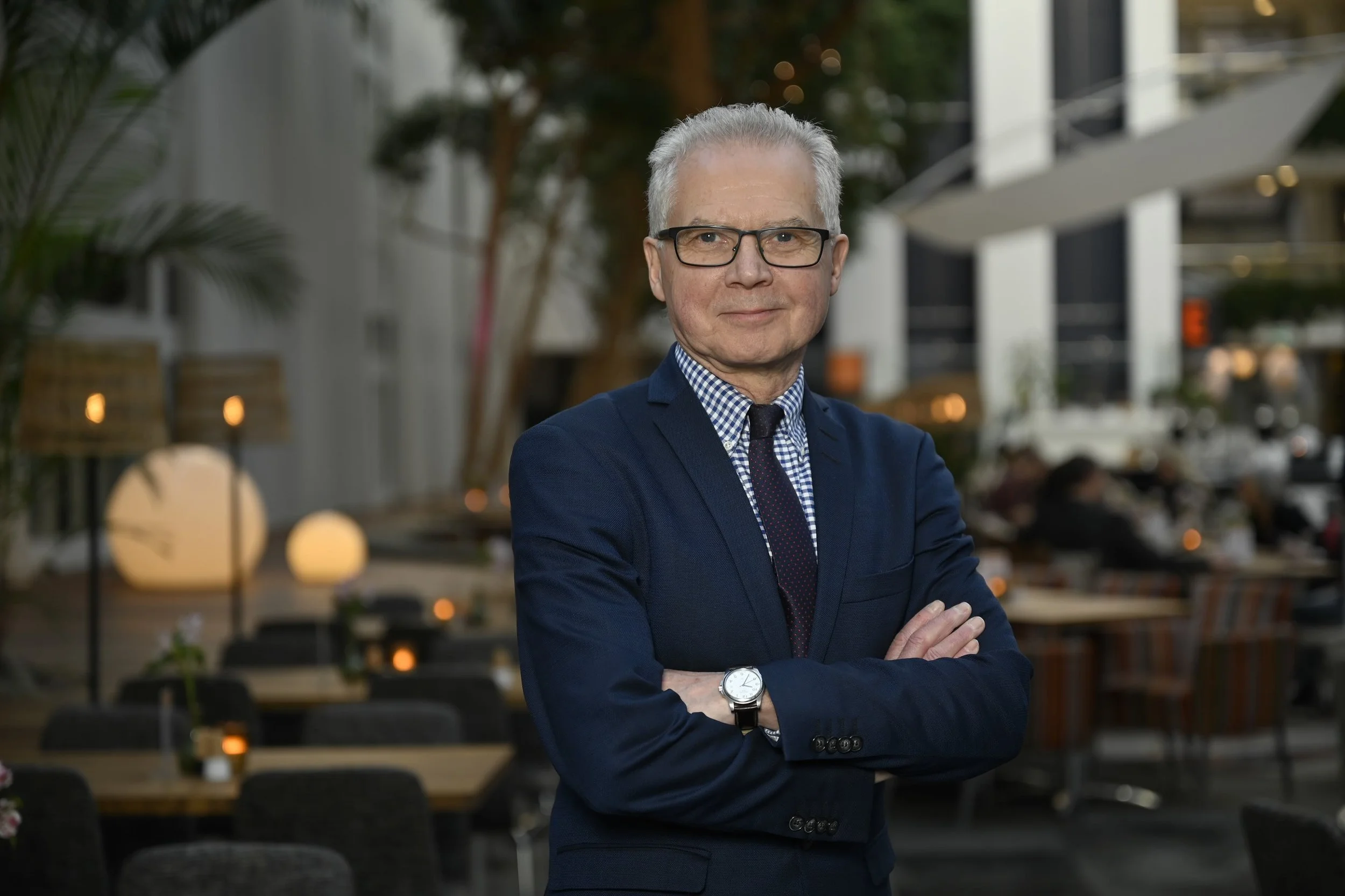 A man with gray hair, glasses, wearing a navy blue suit, checked shirt, and tie, standing with arms crossed in an outdoor café or restaurant setting during evening.