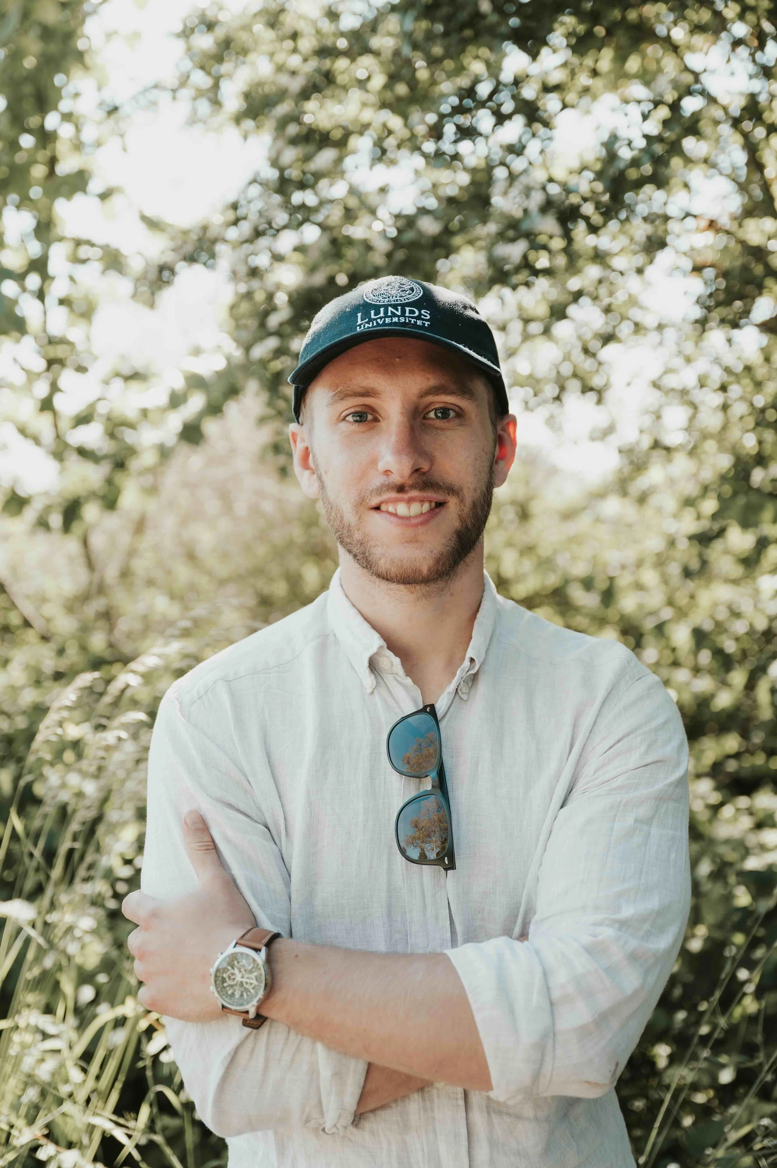A young man with light skin and facial hair, wearing a dark blue cap with the logo and text of Lunds University, stands outdoors among green trees. He is smiling, crossing arms, with sunglasses hanging from his shirt and a wristwatch on his left wrist.