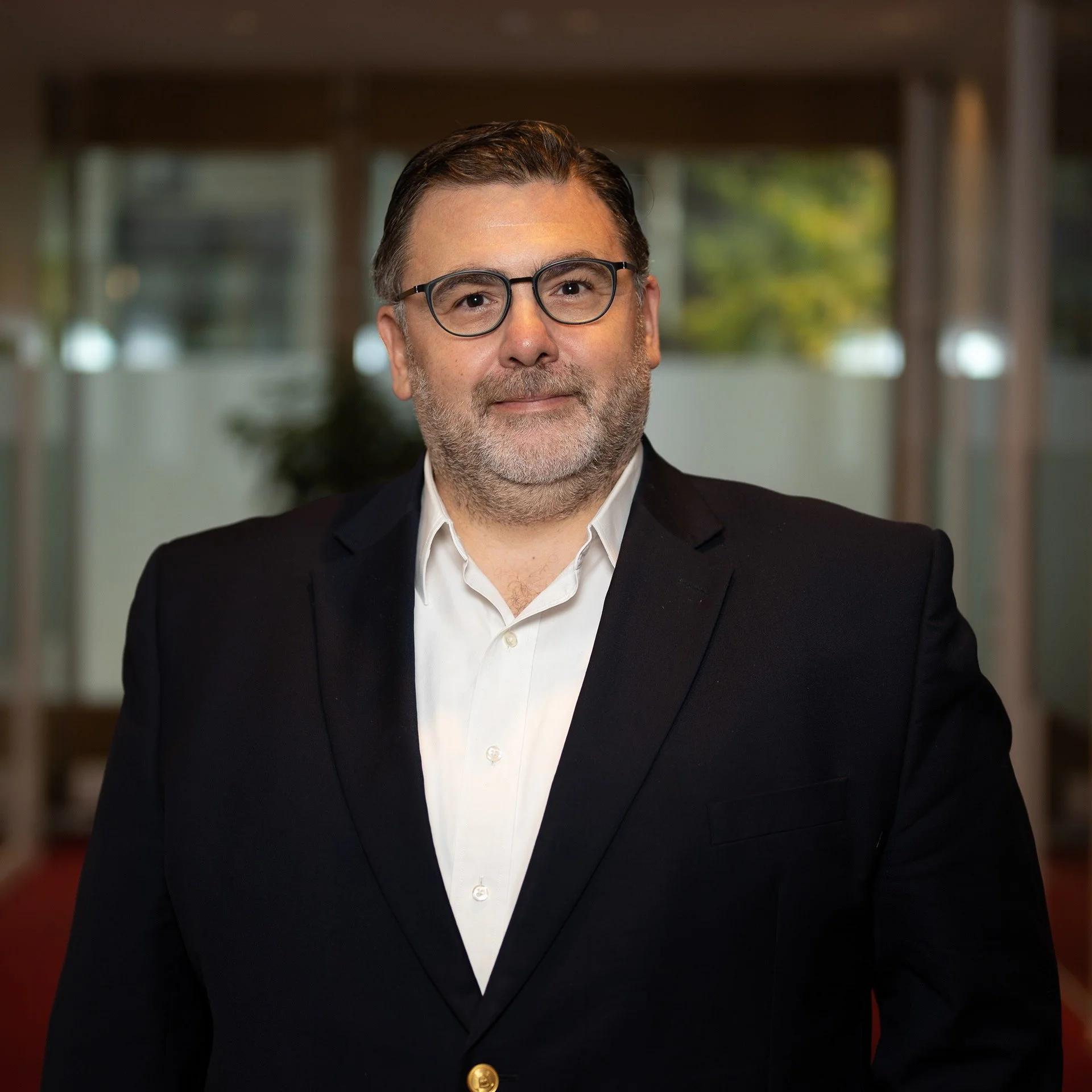 A middle-aged man with glasses, grey beard, in a black suit with a white shirt, standing indoors with a blurred background of windows and greenery.