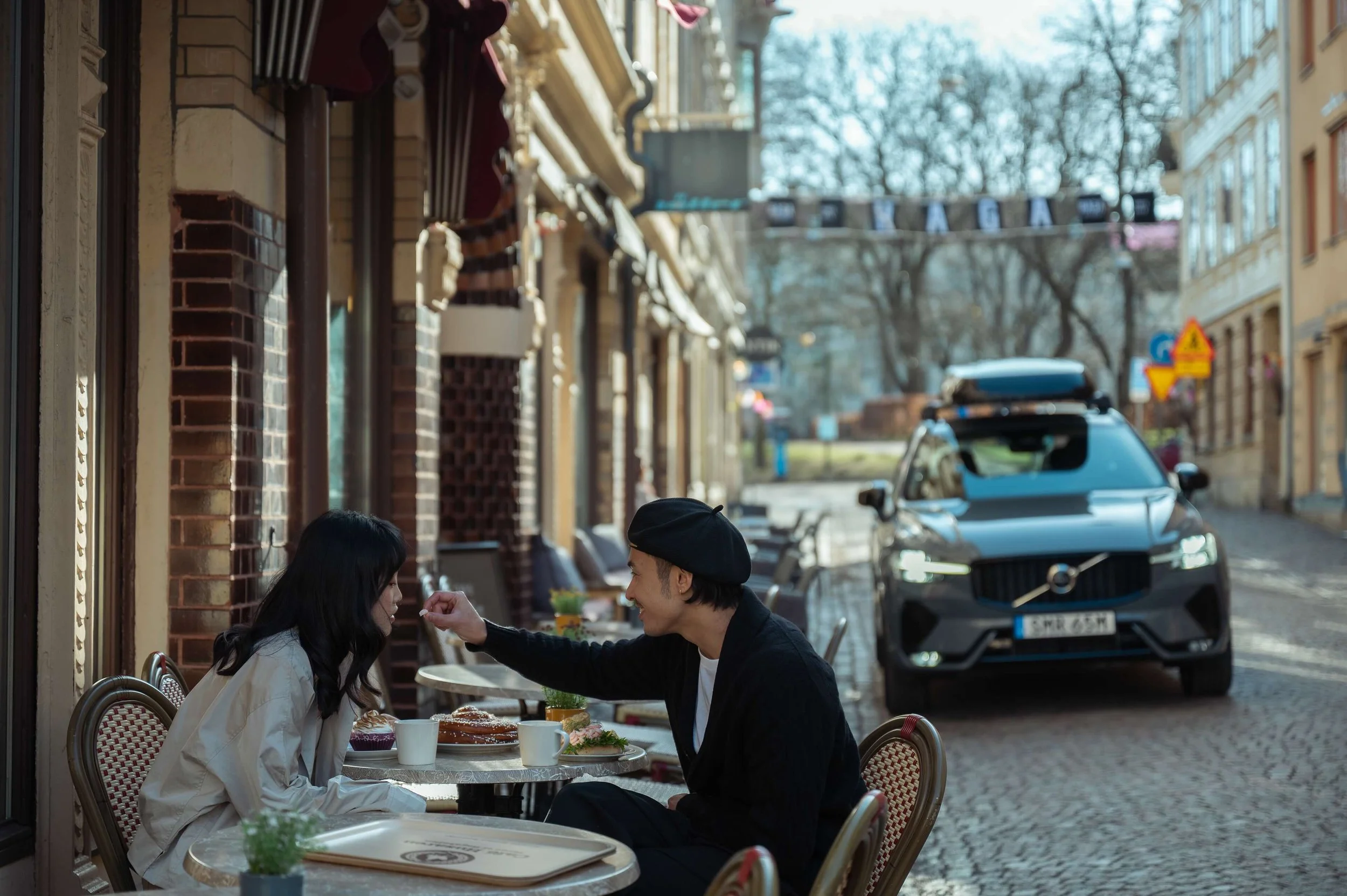 A man feeding a woman at an outdoor café on a cobblestone street, with parked car and buildings in the background.