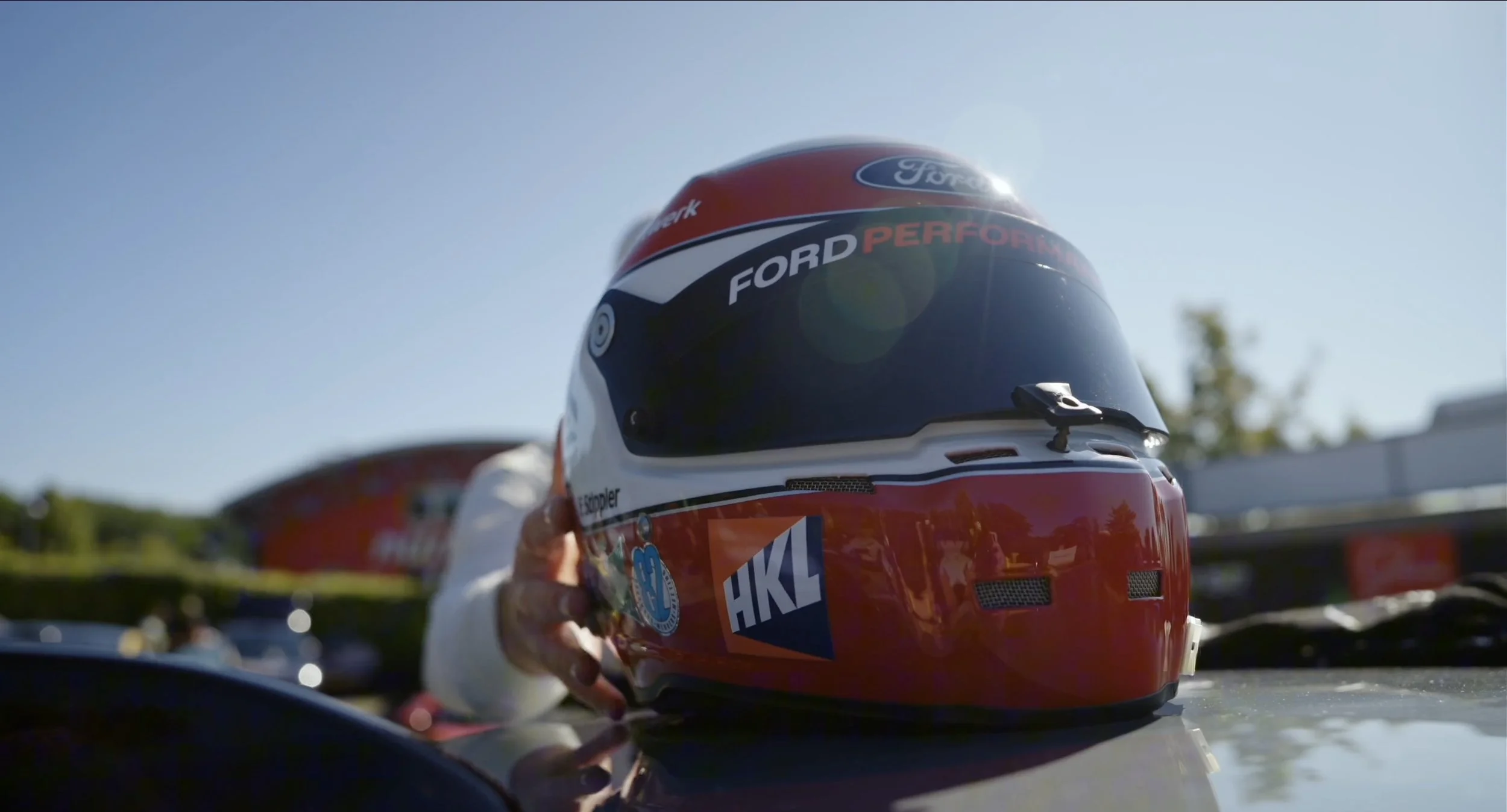 A race car driver holding a red and black racing helmet with sponsorship logos, outdoors on a sunny day.