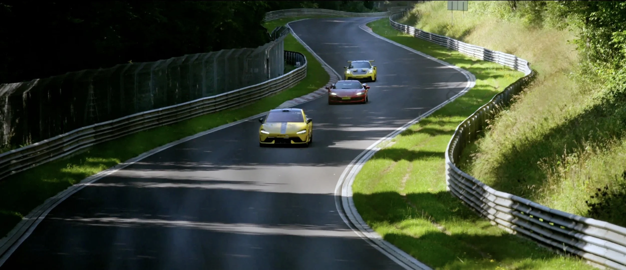 Four sports cars driving on a winding mountain race track surrounded by greenery and metal guardrails, with trees casting shadows on the road.